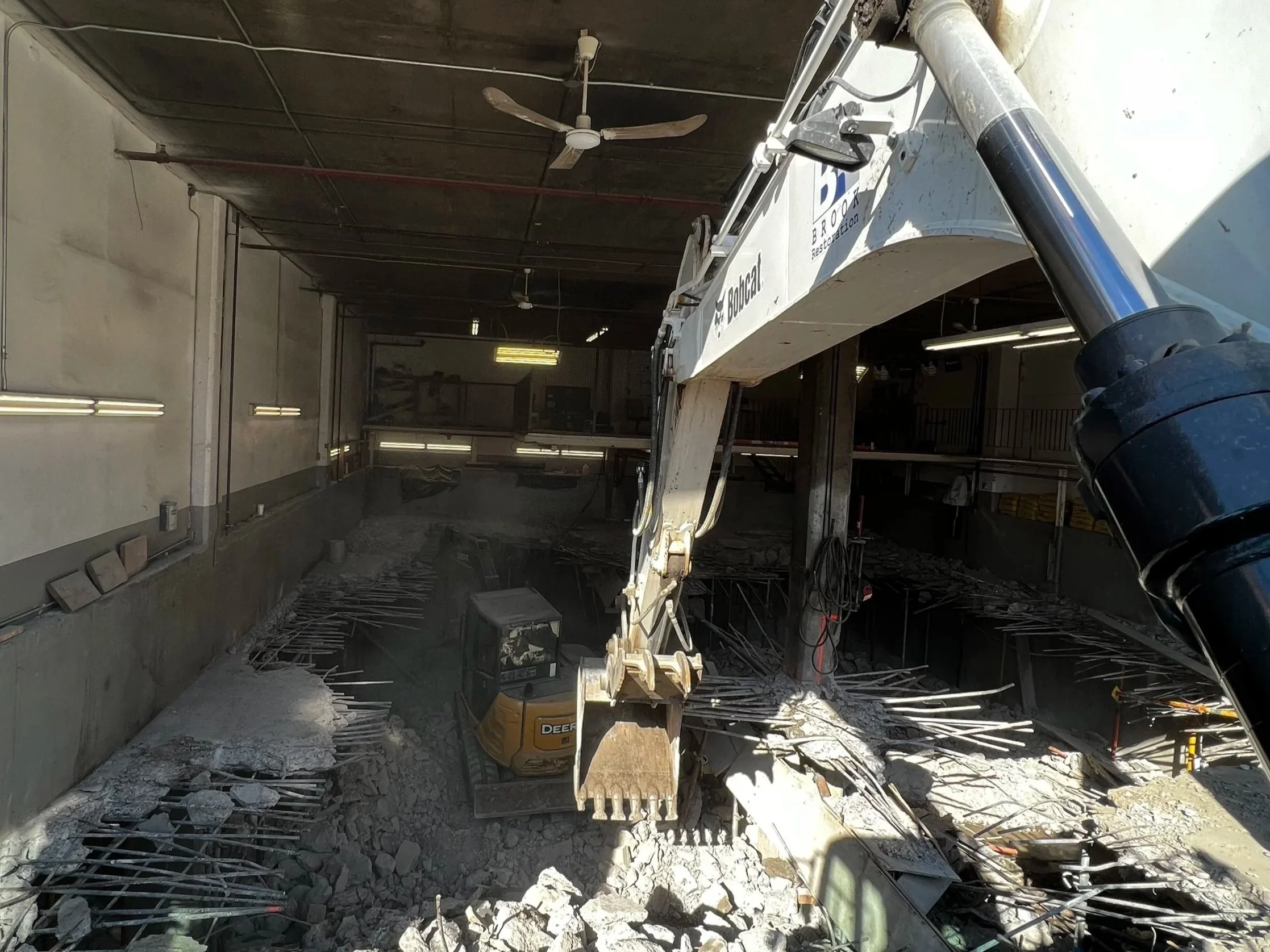 Construction site inside a building with a large excavator demolishing the floor and rubble scattered around.