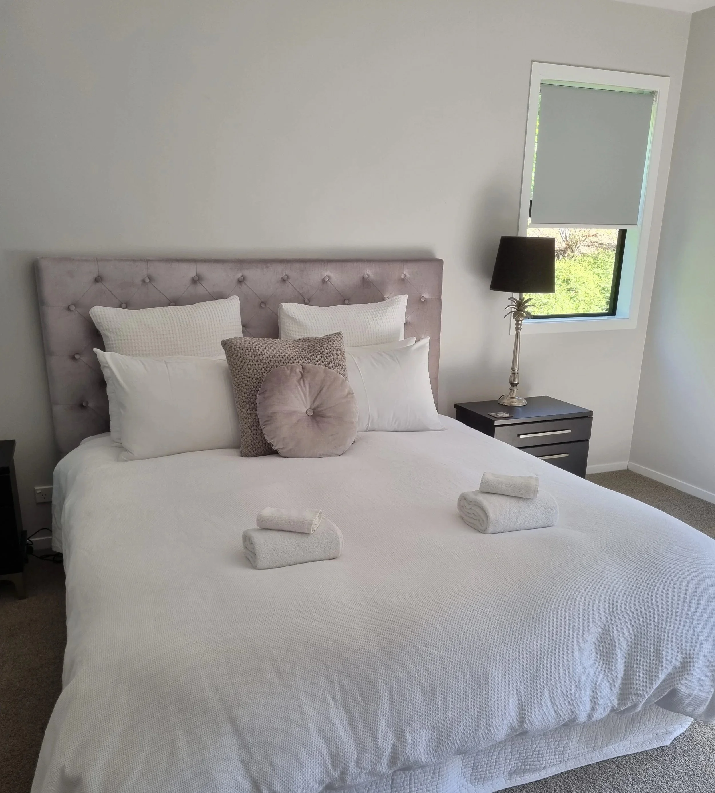 A neatly made bed with white linens, a tufted pink headboard, and six pillows, including ones with textured and smooth covers. Three rolled towels are placed on the bed, and a bedside table with a black lamp and small decorative object is positioned next to a window with partially closed blinds, revealing greenery outside.