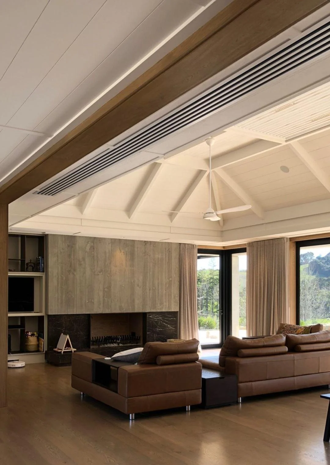 Living room with vaulted ceiling, white beams, ceiling fan, large windows with curtains, brown leather sofas, fireplace, wooden floors, and open view of greenery outside.