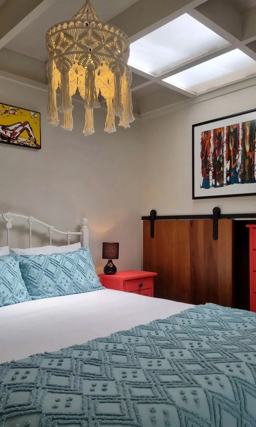 A bedroom with a white bed, light blue textured bedding, and matching pillows. There is a pink nightstand with a small black lamp, a decorative wooden sliding door, and framed artwork on the walls. A woven white chandelier hangs from the ceiling, and skylights provide natural light.