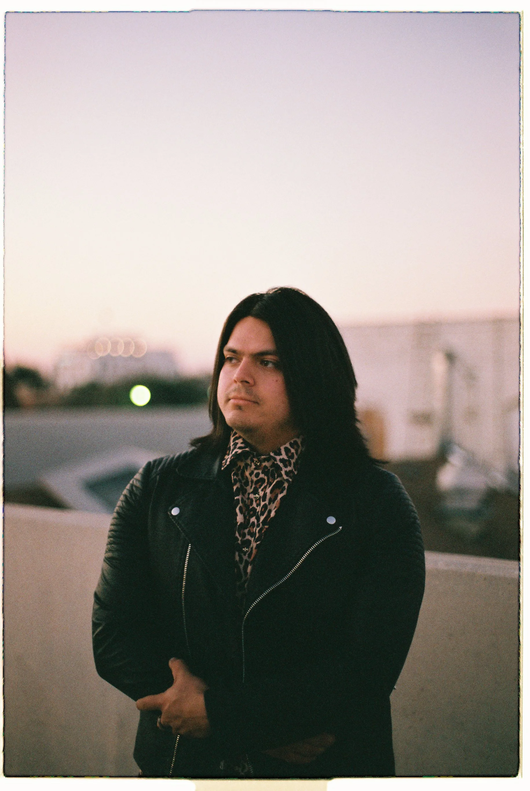 Portrait of a person with long dark hair wearing a black jacket and leopard-print shirt outdoors at dusk
