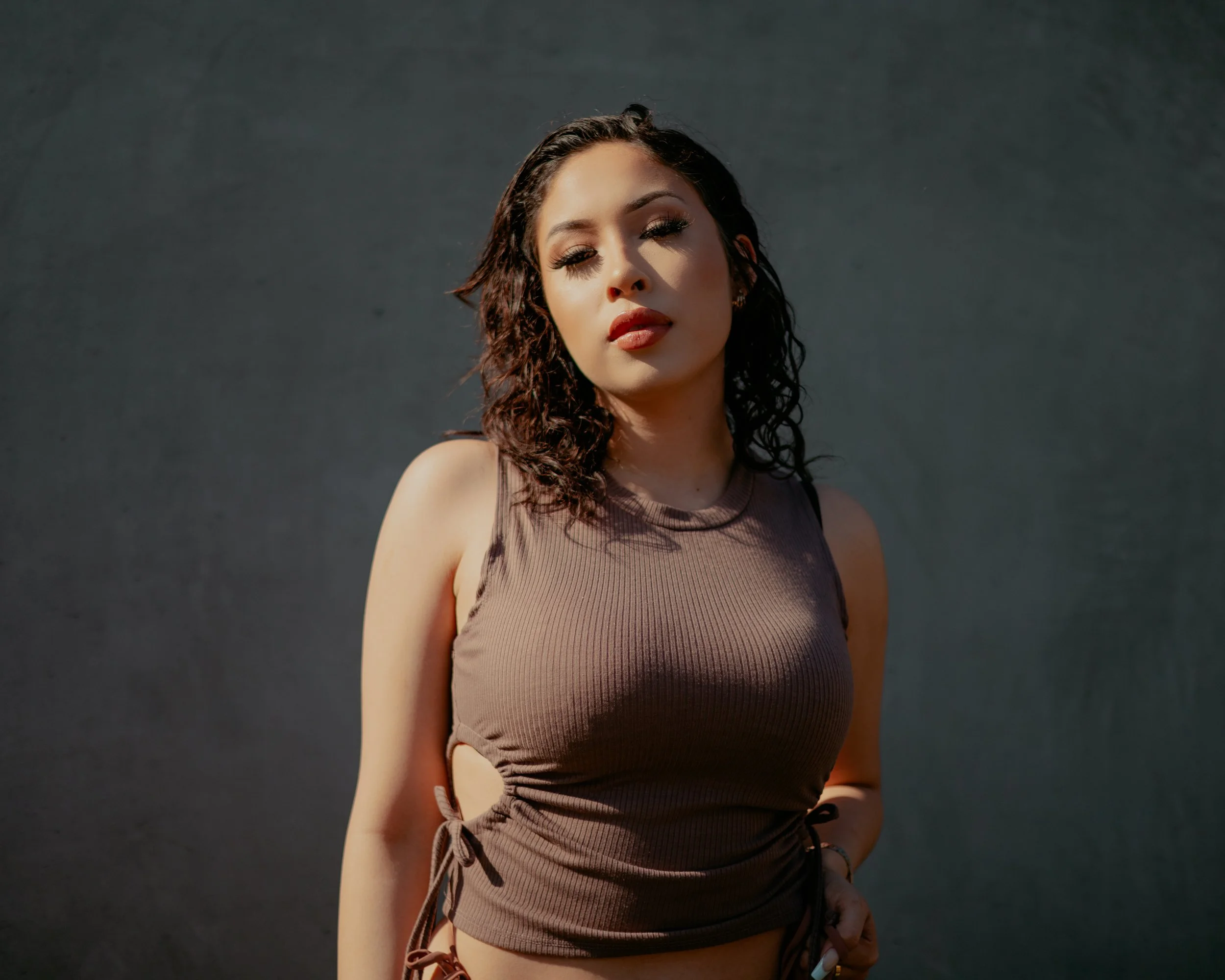 Portrait of a woman with curly dark hair wearing a brown ribbed sleeveless top against a dark wall