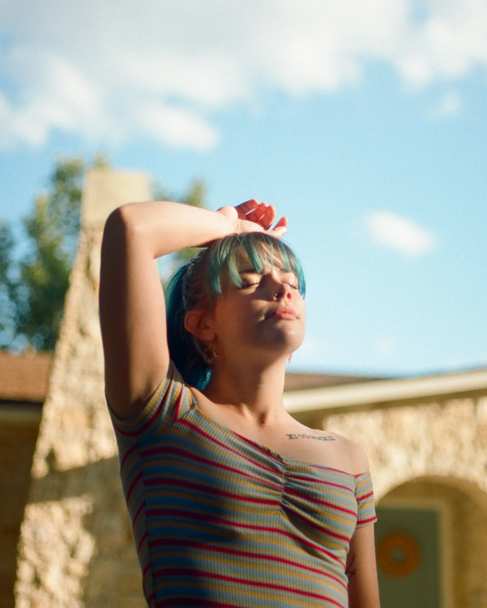 Young woman with teal hair stretching outdoors in bright sunlight, wearing a striped shirt against a blue sky.