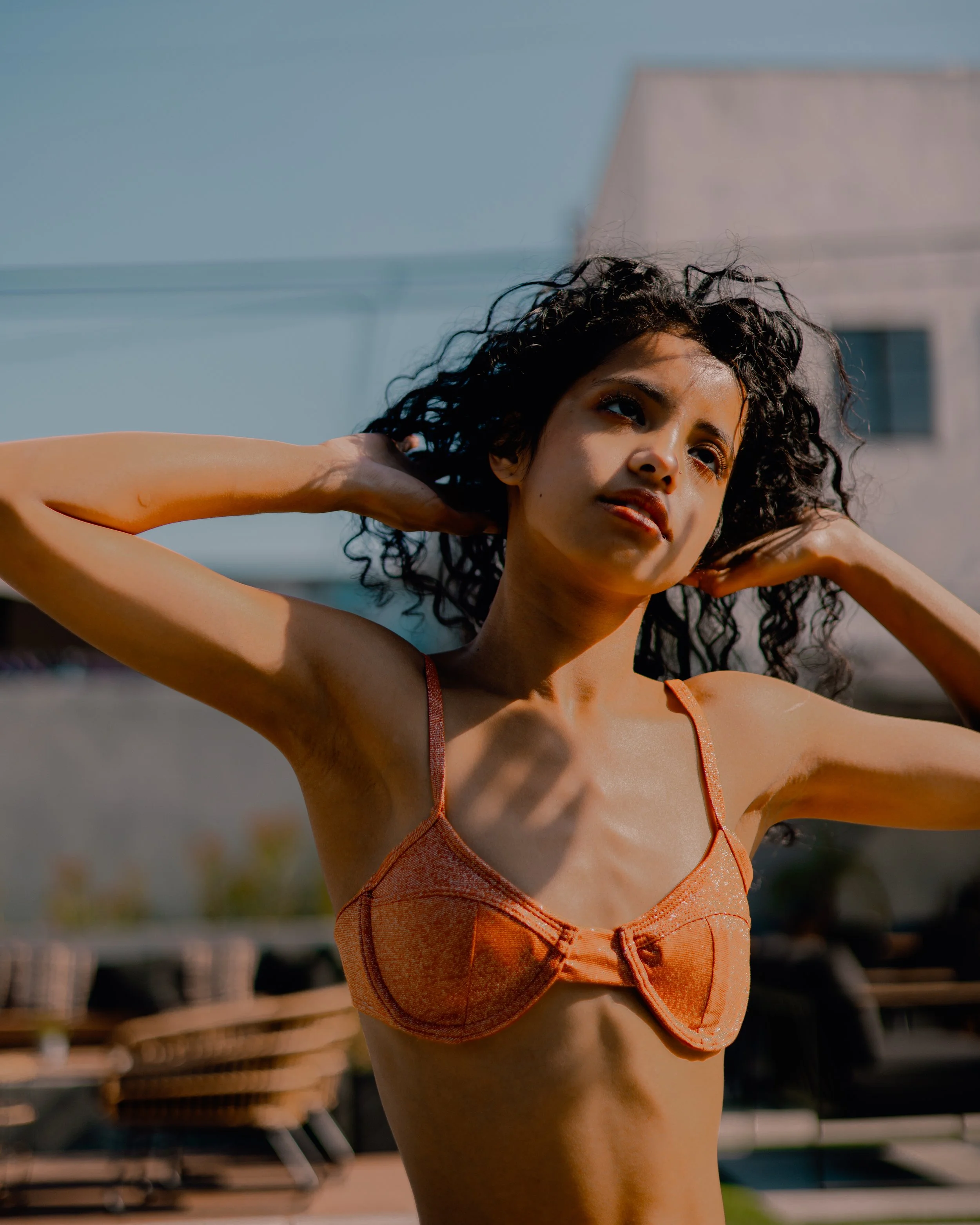 Close-up portrait of a curly-haired woman in an orange bikini posing outdoors by a pool.