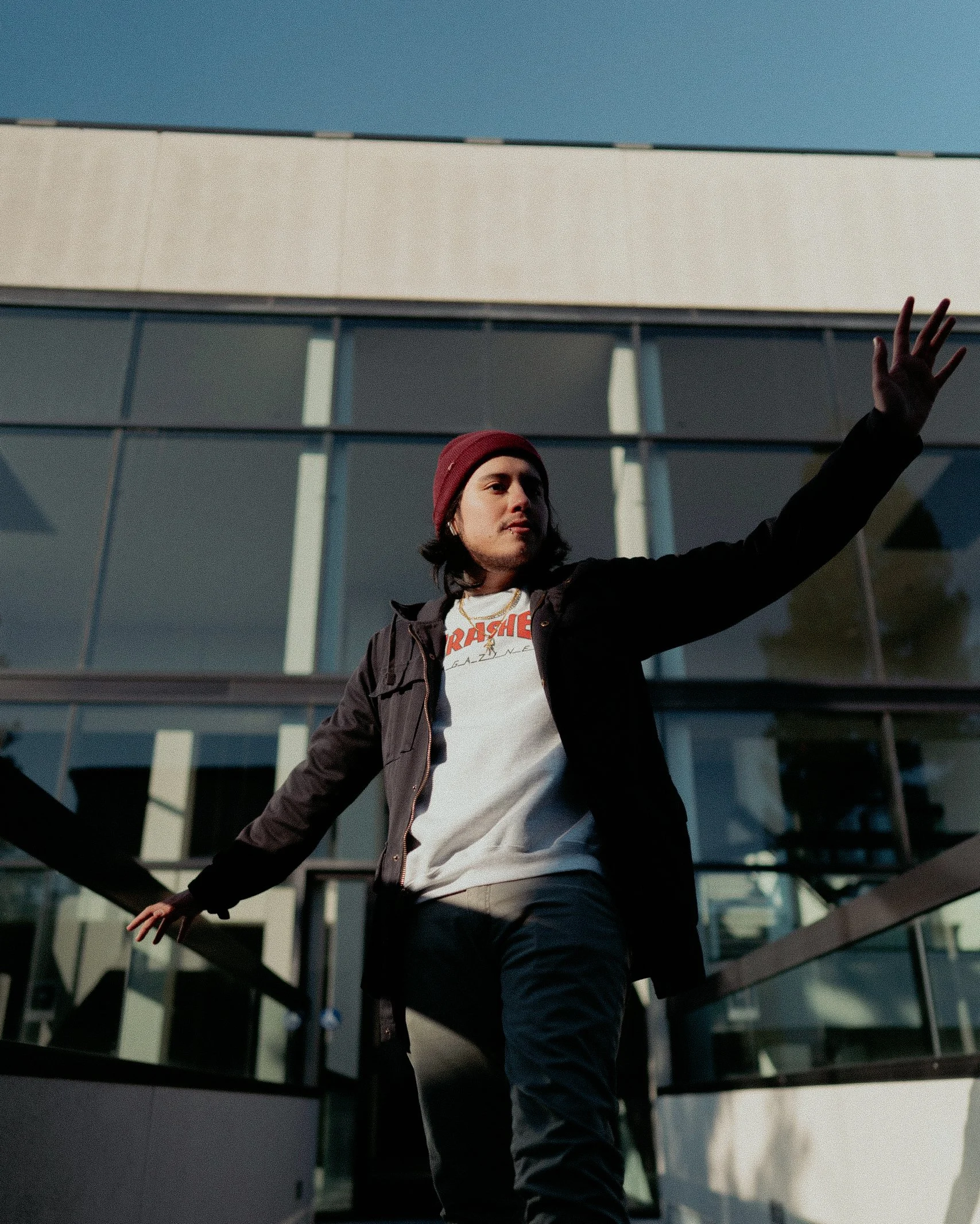 Person in a red beanie and black jacket standing outdoors with arms outstretched in front of a modern glass building