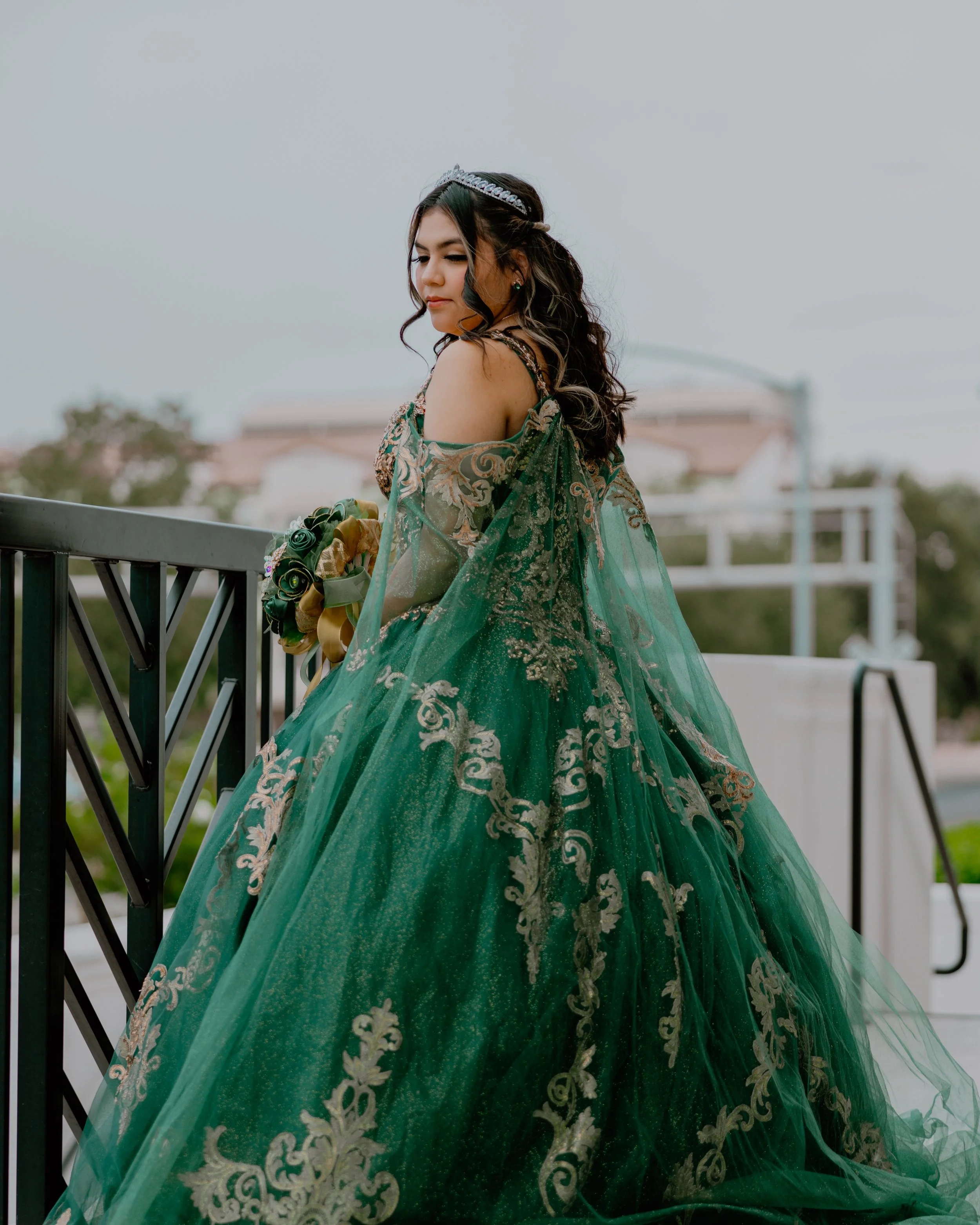 Woman in an ornate green embellished gown and tiara holding a bouquet on a balcony