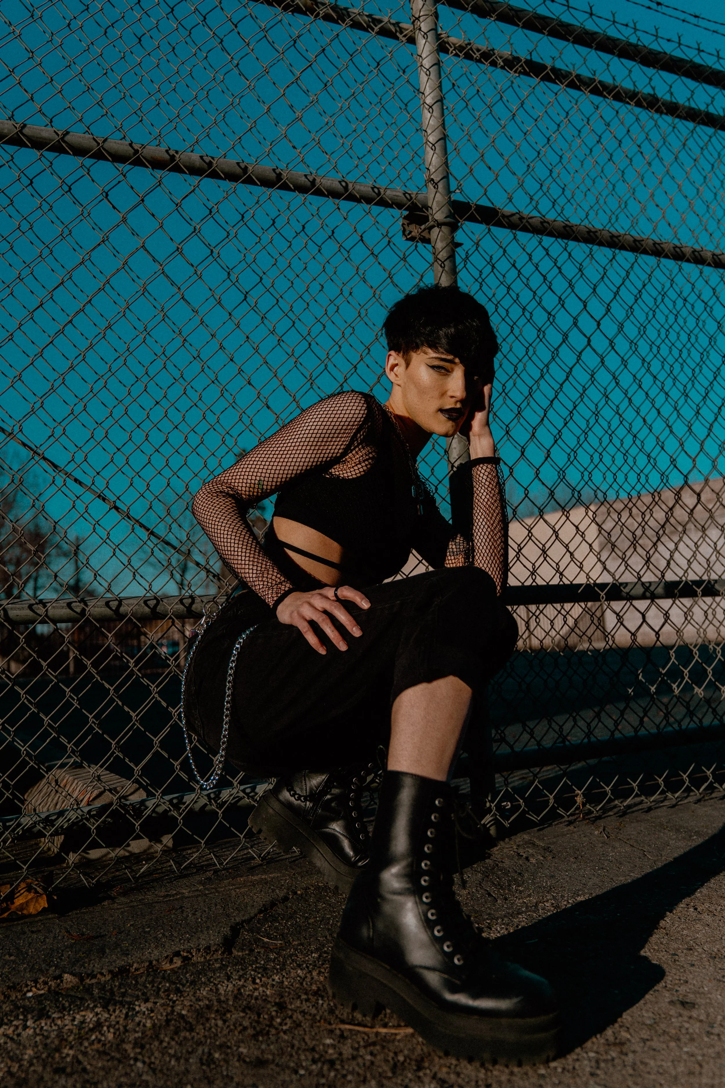 Fashion portrait of a person in black fishnet sleeves and lace-up boots sitting against a chain-link fence under a blue sky