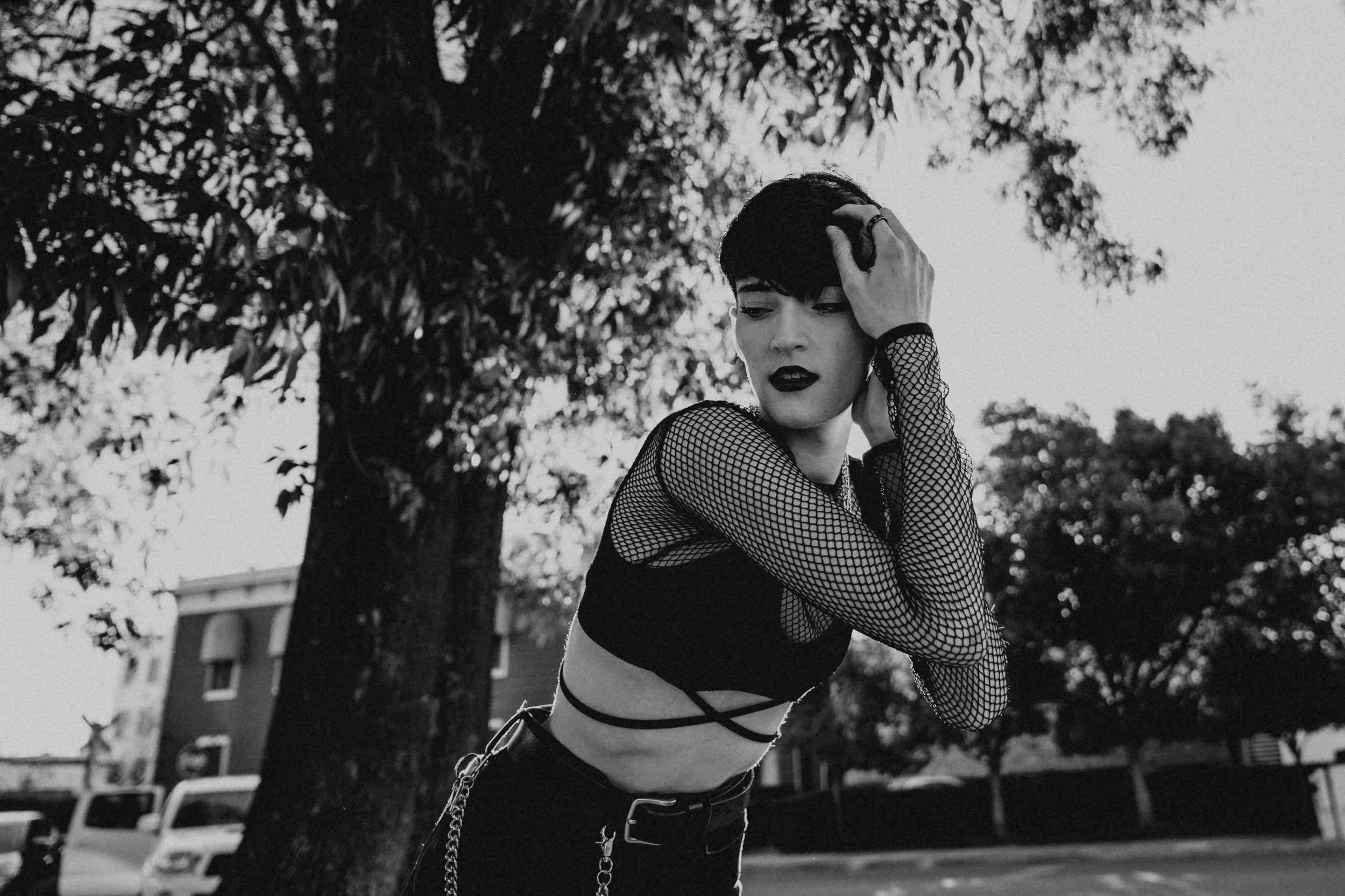 Black-and-white portrait of a woman in fishnet sleeves and dark clothing posing outdoors beneath trees.