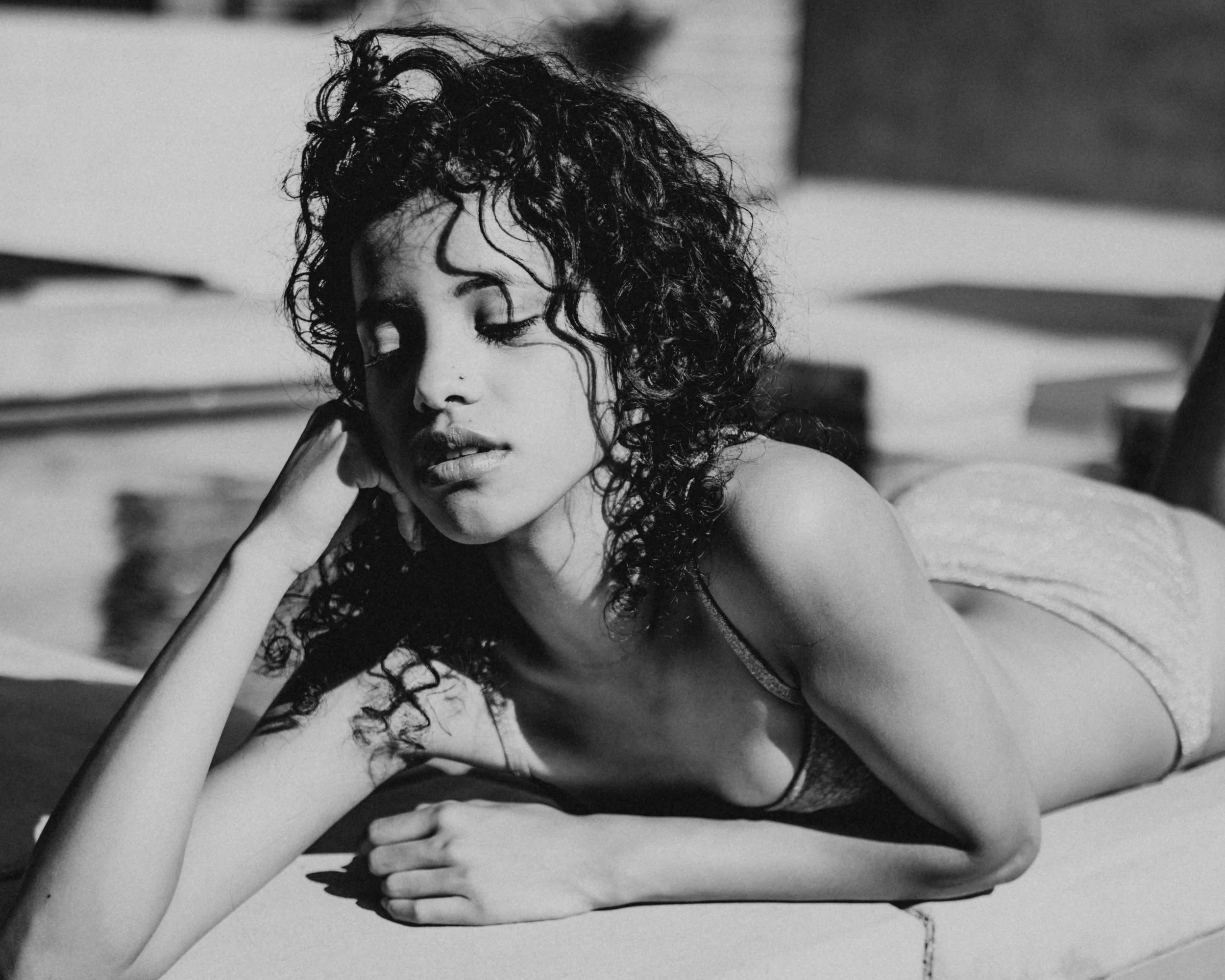 Black-and-white portrait of a curly-haired woman reclining beside a pool with her head resting on her hand.