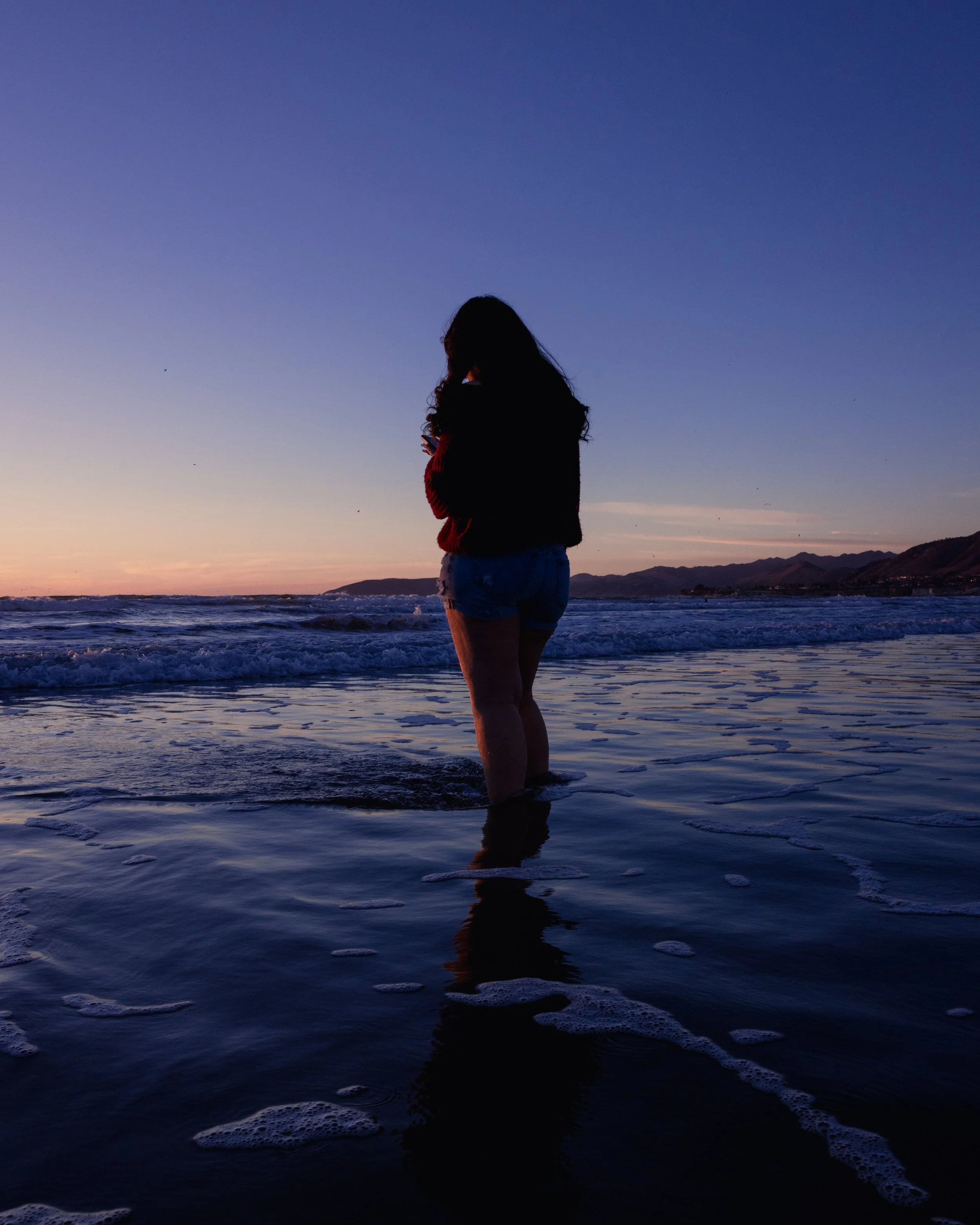 Silhouette of a woman standing in ocean waves at sunset with mountains in the background