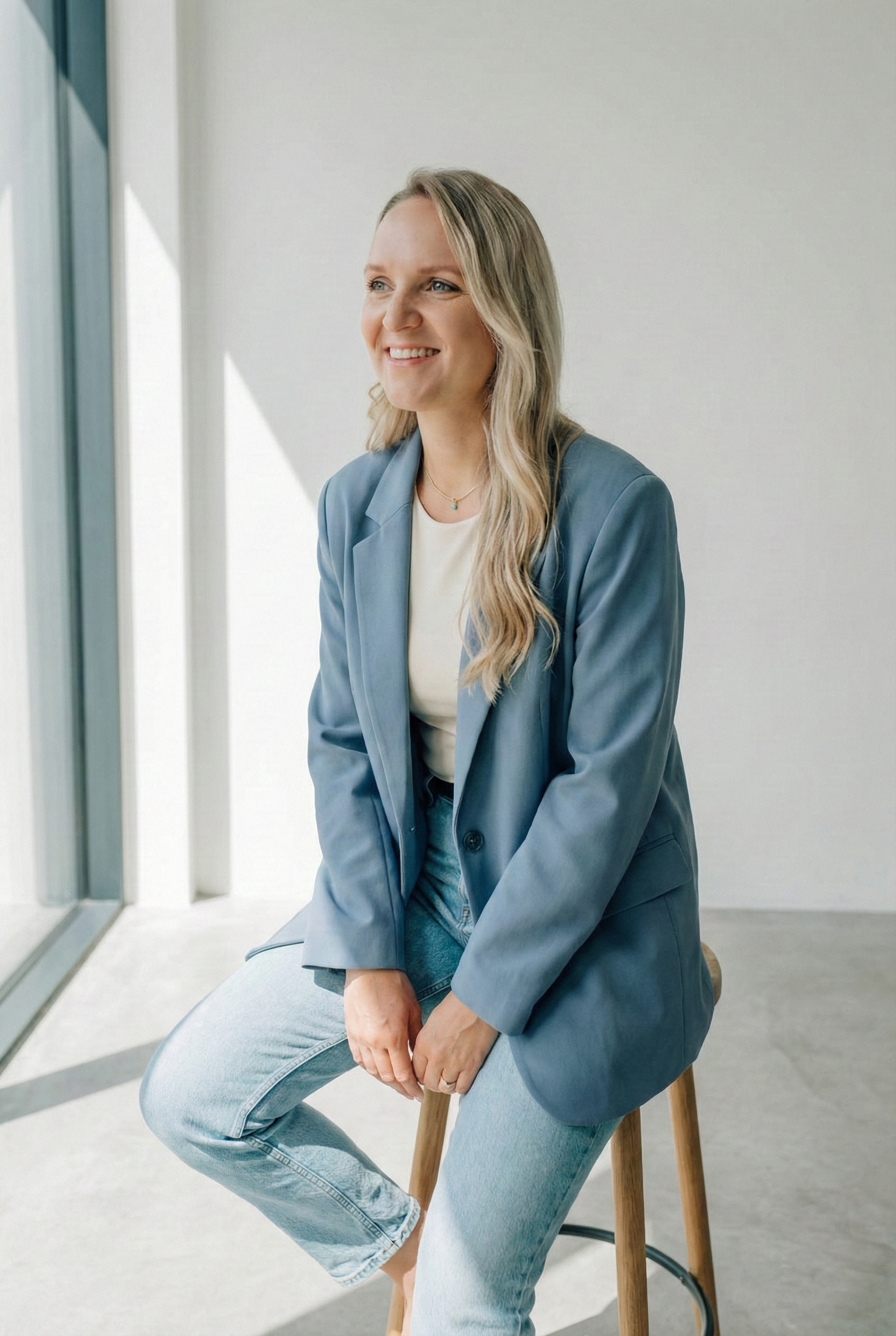 Eine Frau sitzt auf einem Barhocker in einem hellen Raum, trägt eine blaue Jacke, ein weißes Top und Jeans, lächelt und schaut nach draußen.