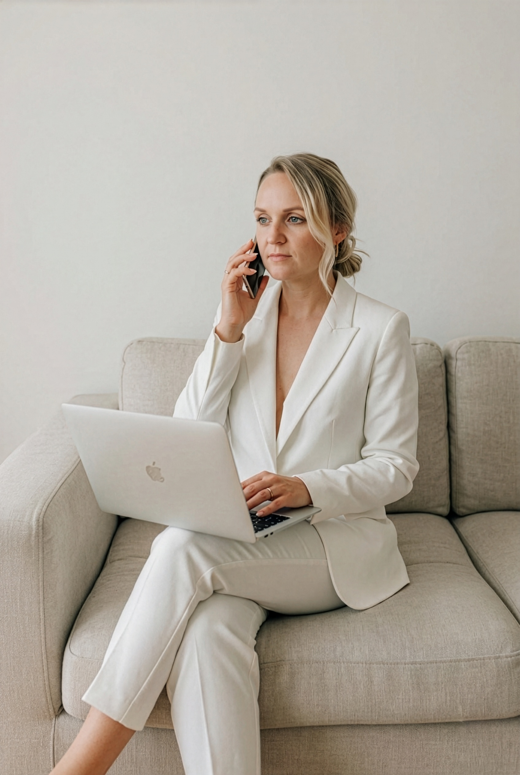 Frau in weißem Anzug sitzt auf einem Sofa, spricht am Telefon und benutzt einen Laptop.