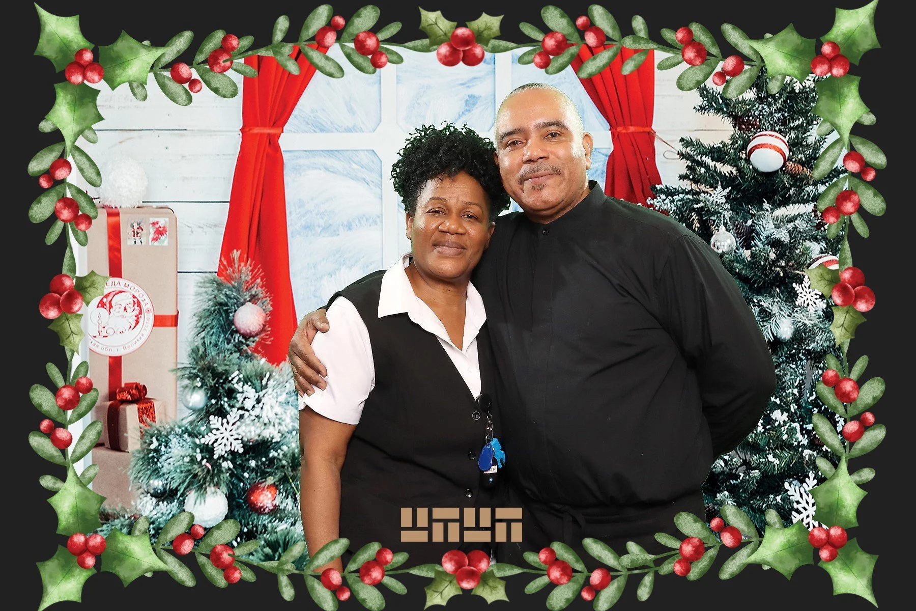 A couple standing together in front of a Christmas decorated background, including a Christmas tree with ornaments, wrapped presents, and red curtains, with a holiday-themed border of holly leaves and red berries surrounding the image.