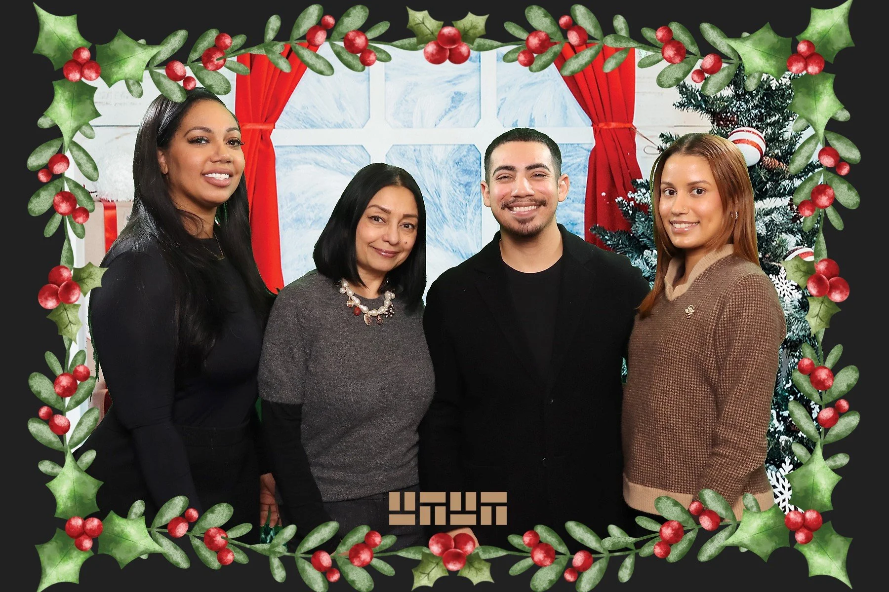 Four people standing in front of a snowy window with red curtains and a decorated Christmas tree, framed by a green holly and red berry border.