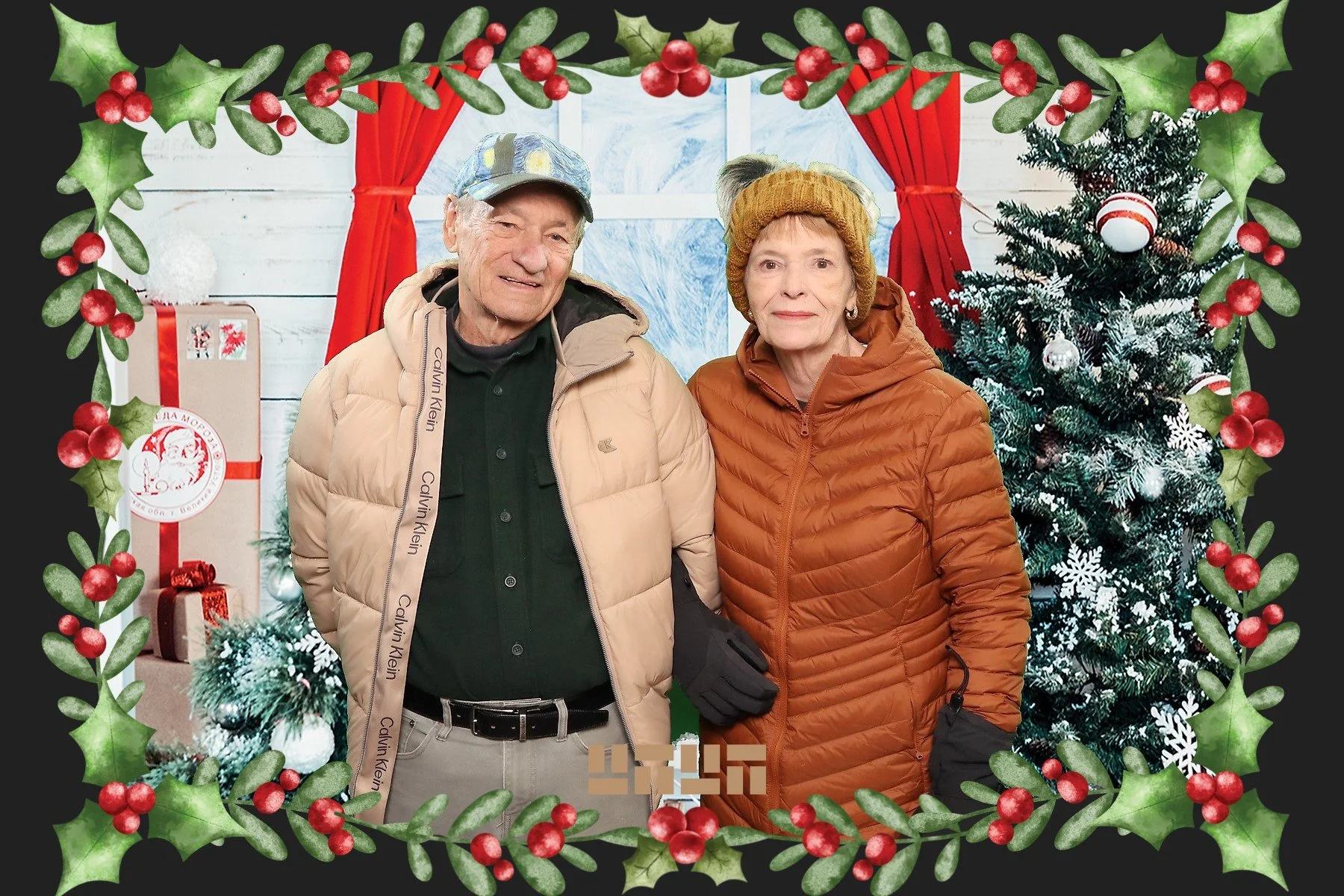 An elderly man and woman dressed in winter clothing standing inside a Christmas decorated room. A Christmas tree with ornaments is visible to the right, and wrapped presents are seen behind them. The scene is framed with a festive border of holly wit