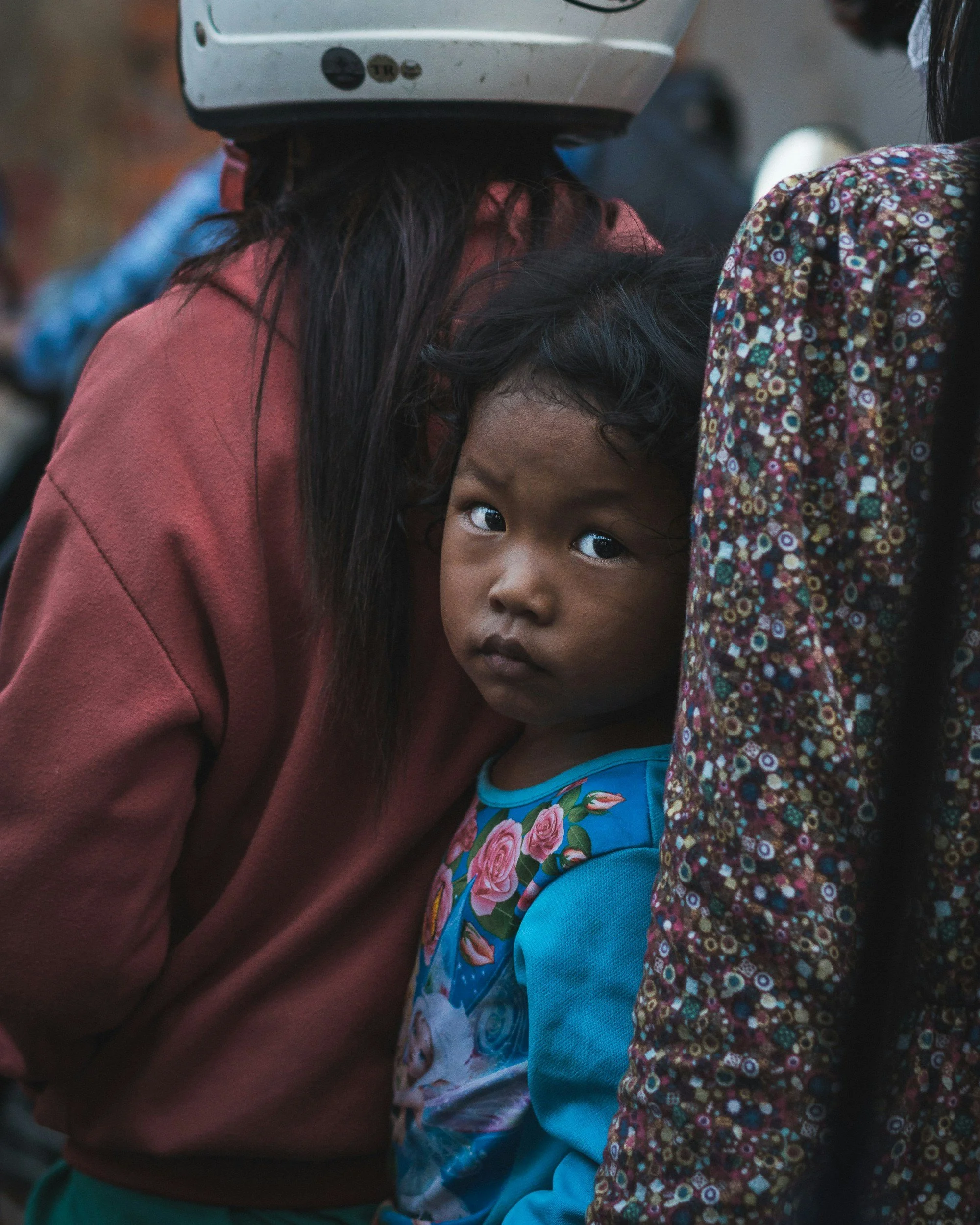 A young girl with curly black hair and dark eyes looking directly at the camera, wearing a colorful dress with roses. An adult wearing a maroon jacket and a helmet is leaning over her, with the adult's hair and part of their face visible.
