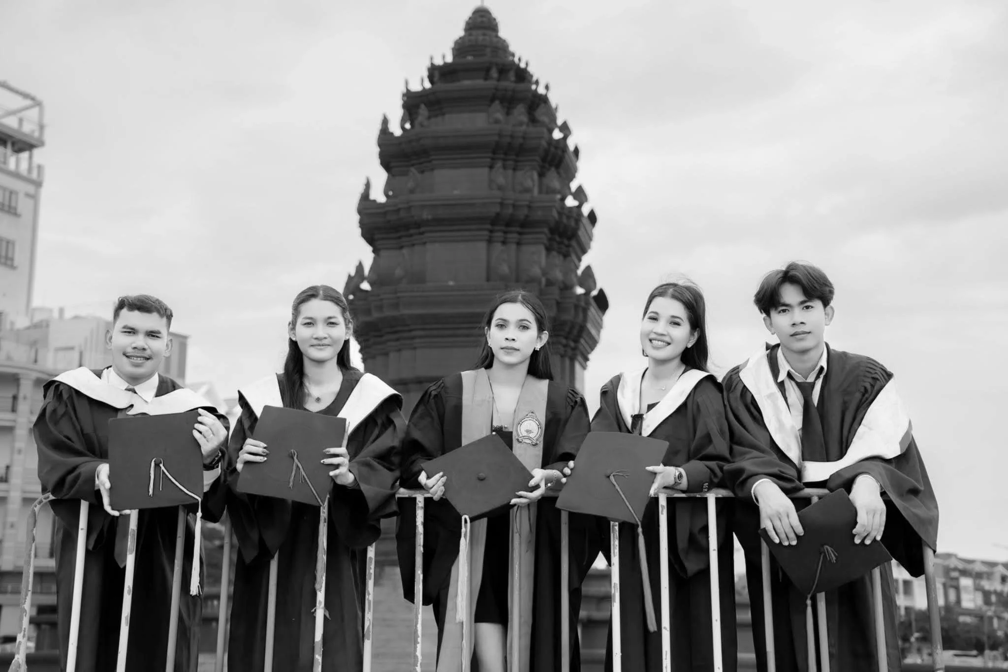 Five graduates in caps and gowns standing behind a metal barricade in front of a historic tower with a domed roof.