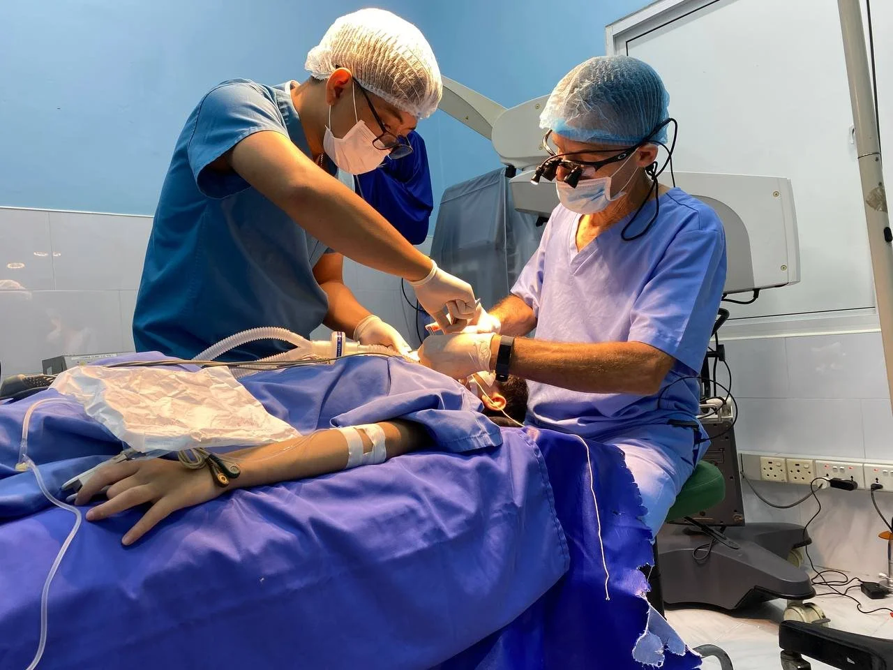 Two surgeons performing surgery in an operating room, with a patient lying on the operating table, surrounded by medical equipment and monitors.