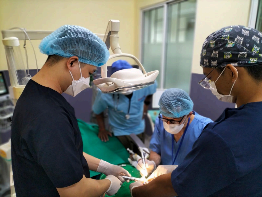 Medical team performing surgery in an operating room with surgeons wearing scrubs, masks, and surgical caps.