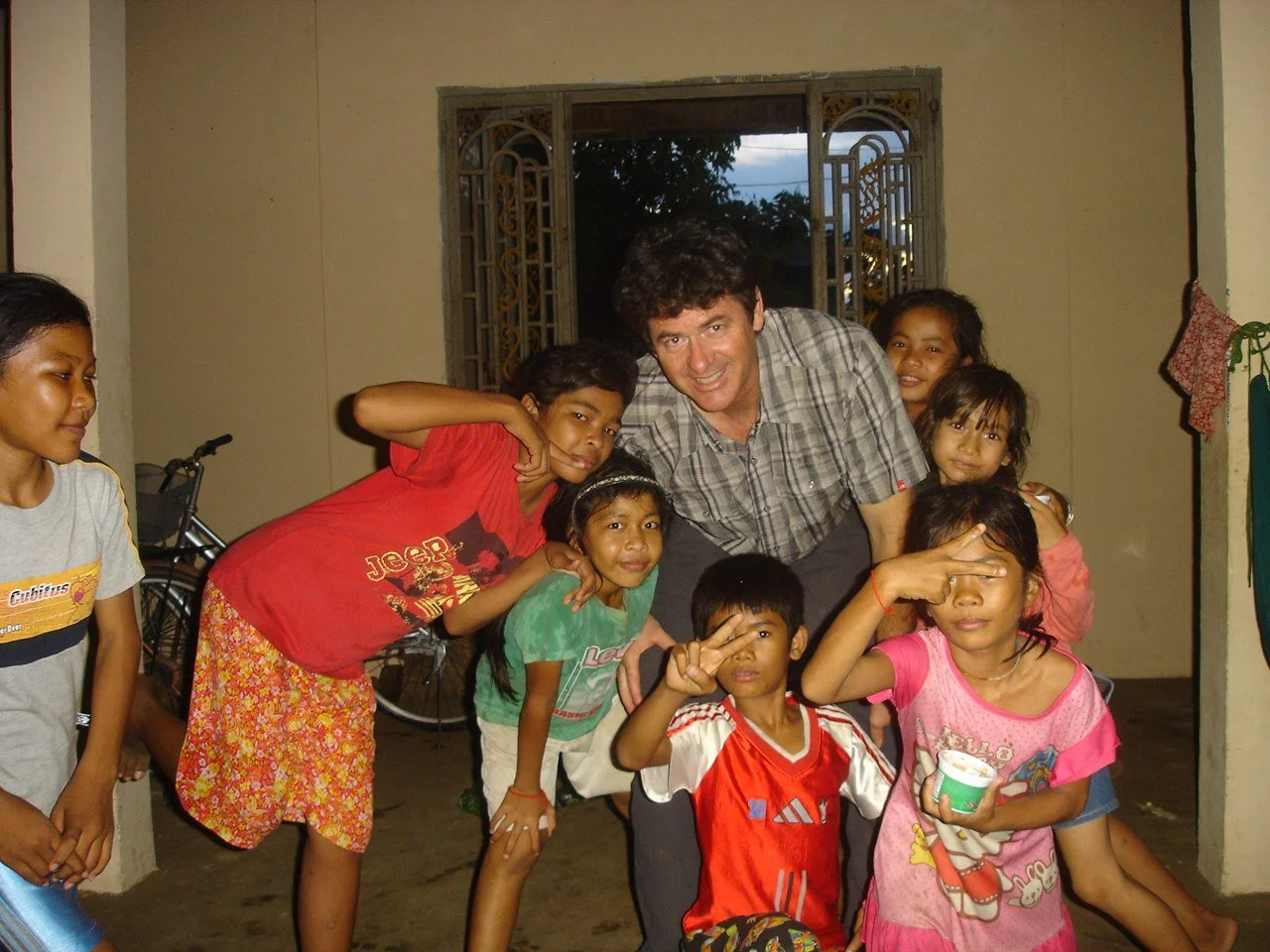 Group of children and a man posing indoors with a window in the background. The children are making various hand gestures and smiling.