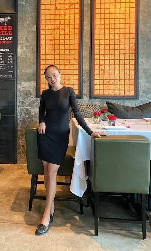 A woman in a black dress and heels standing in a restaurant next to a table with a white tablecloth and green chairs, smiling at the camera.
