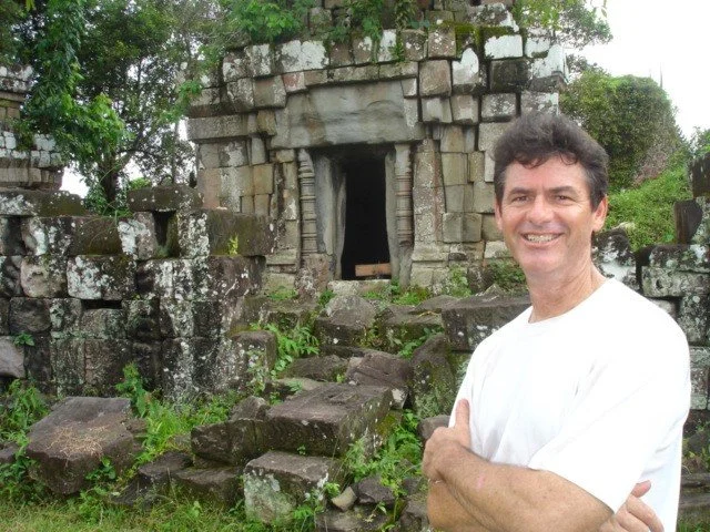 A man standing with crossed arms smiling in front of ancient stone ruins with a dark doorway, surrounded by greenery.