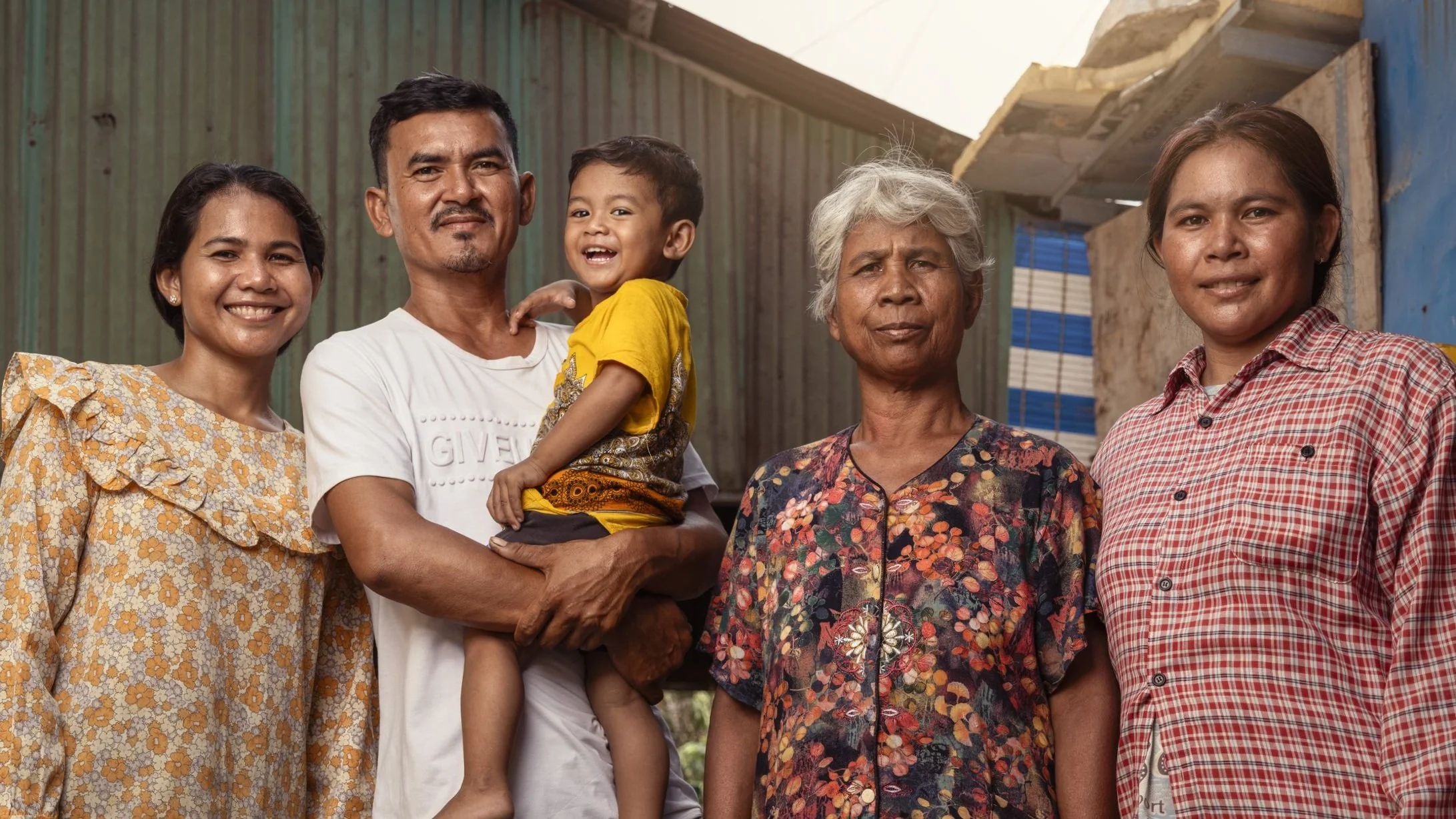 Group of five people, three women, one man, and a young boy, standing outdoors with a rustic, corrugated metal and wood building in the background. They are smiling and appear happy.