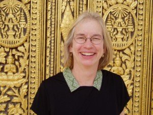 Smiling woman with glasses and blonde hair standing in front of a gilded, ornate door with intricate designs.