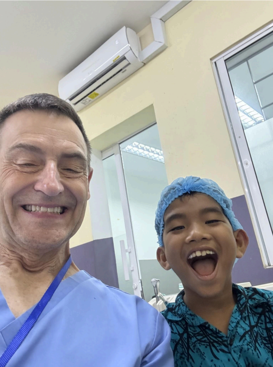 A smiling man and boy take a selfie in a hospital or medical setting. The man is wearing scrubs and the boy is wearing a hair cap, indicating they are in a medical environment.