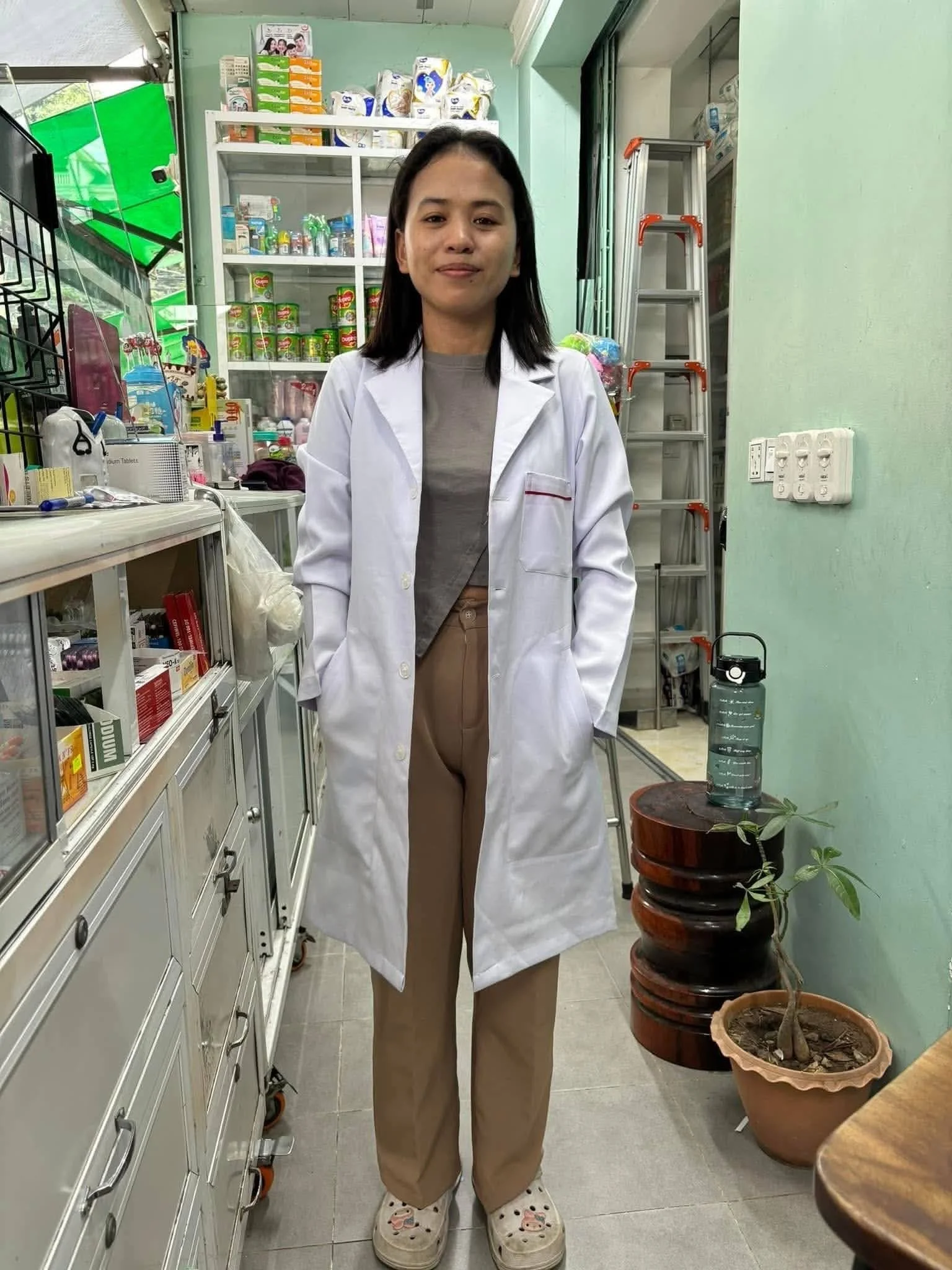 A young woman wearing a white coat and beige pants stands indoors in front of kitchen shelves filled with various food products and cleaning supplies. She is smiling softly and wearing polka dot slippers. A metal ladder is visible behind her, and a potted plant is on her right.