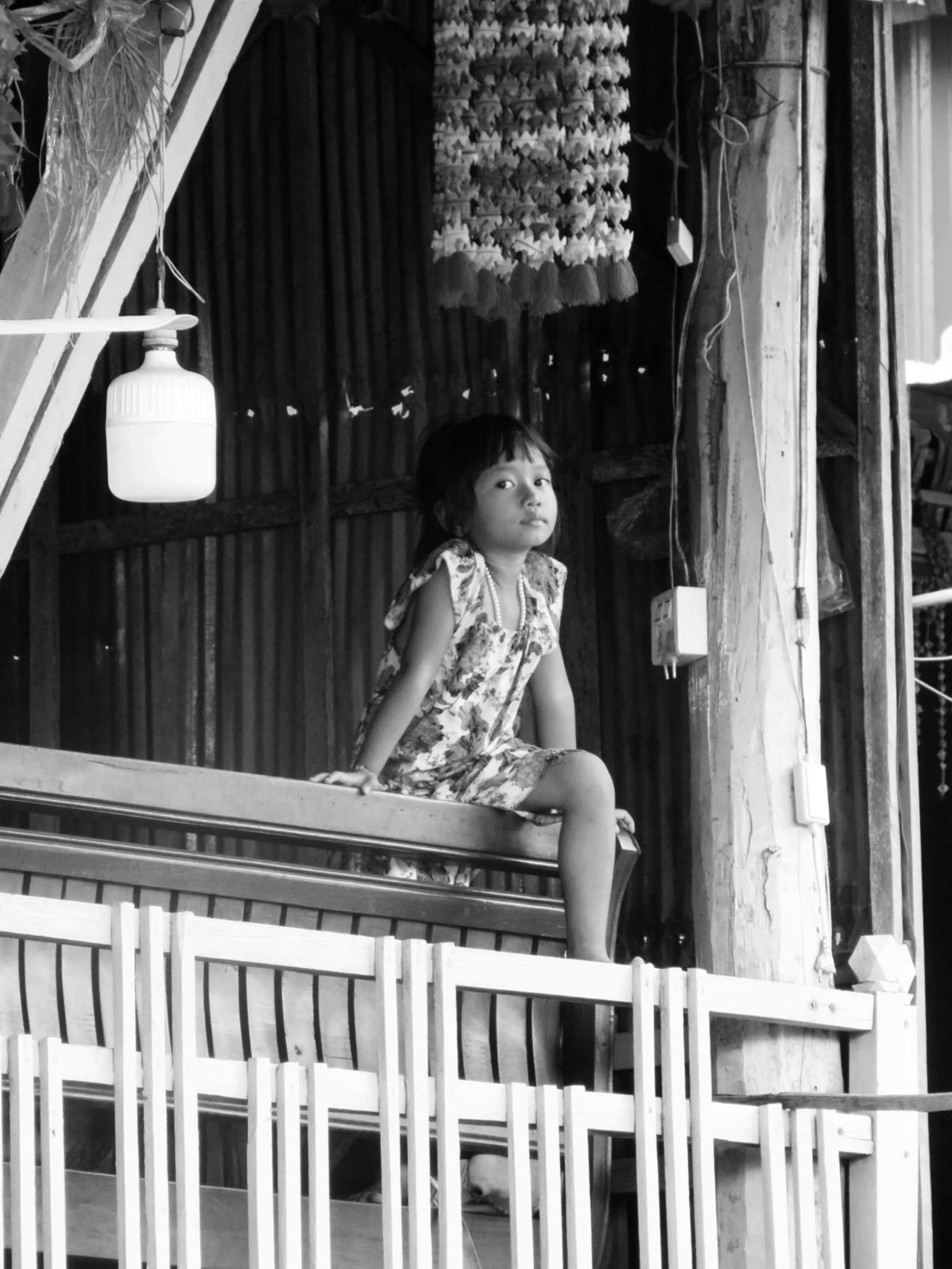 A young girl with short dark hair, wearing a patterned dress, sitting on a wooden balcony or porch with a railing. The background shows a curtain, some hanging decorations, and a wooden wall with vertical planks.