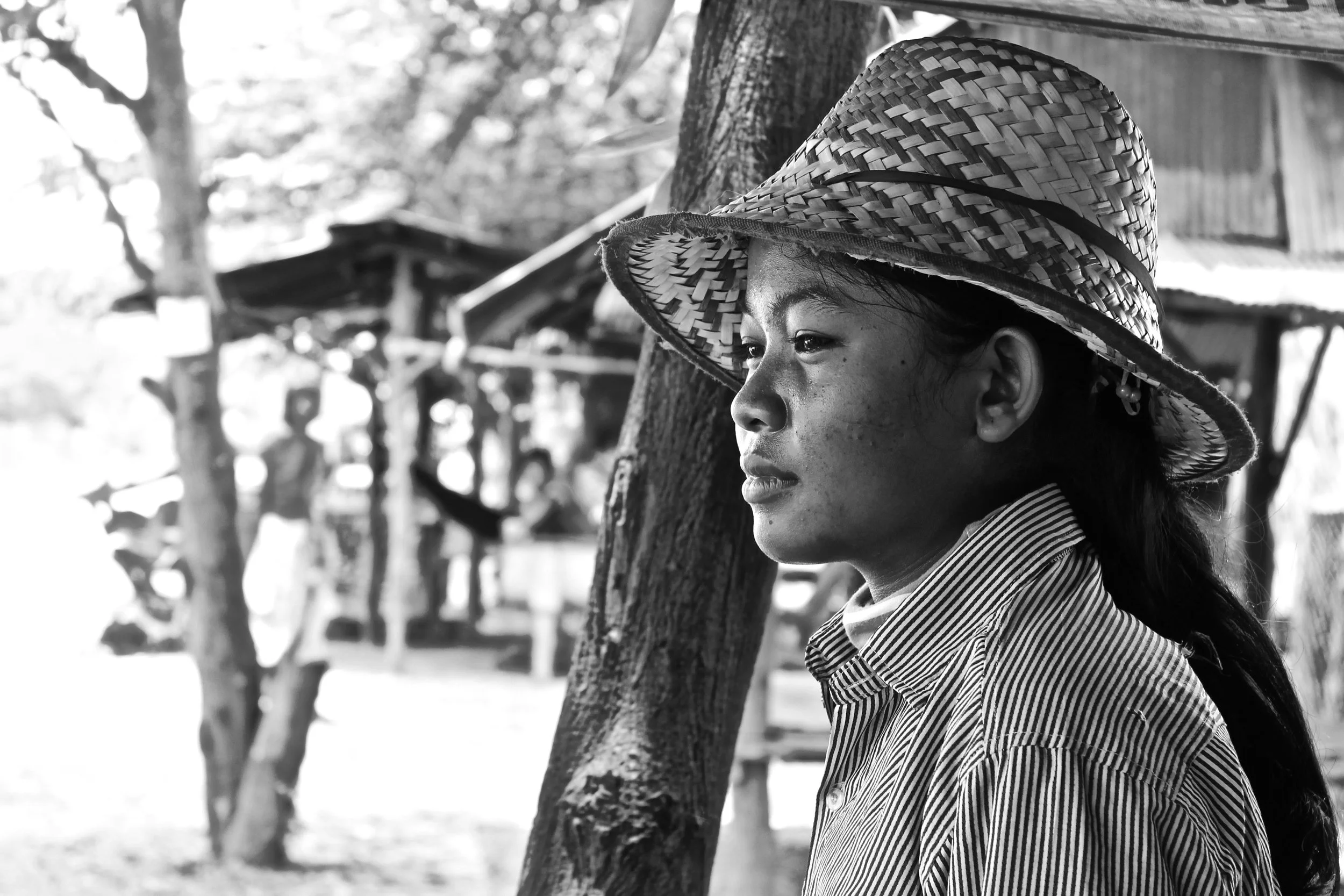 A woman with long hair wearing a striped shirt and large woven hat, standing outdoors near a tree with a blurred background of trees and structures.