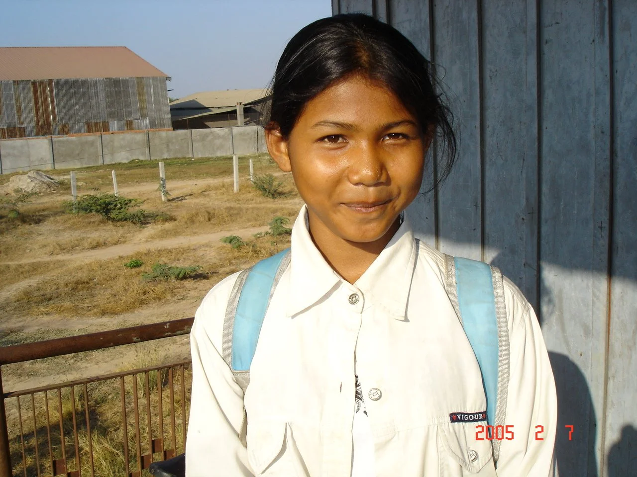 A young girl with dark hair and a light-colored shirt smiling outdoors, carrying a blue backpack.