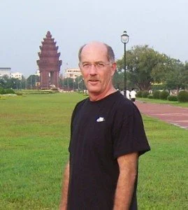 A man with glasses and a black Nike t-shirt standing outdoors on a grassy area, with a large temple structure in the background and a pathway leading up to it.