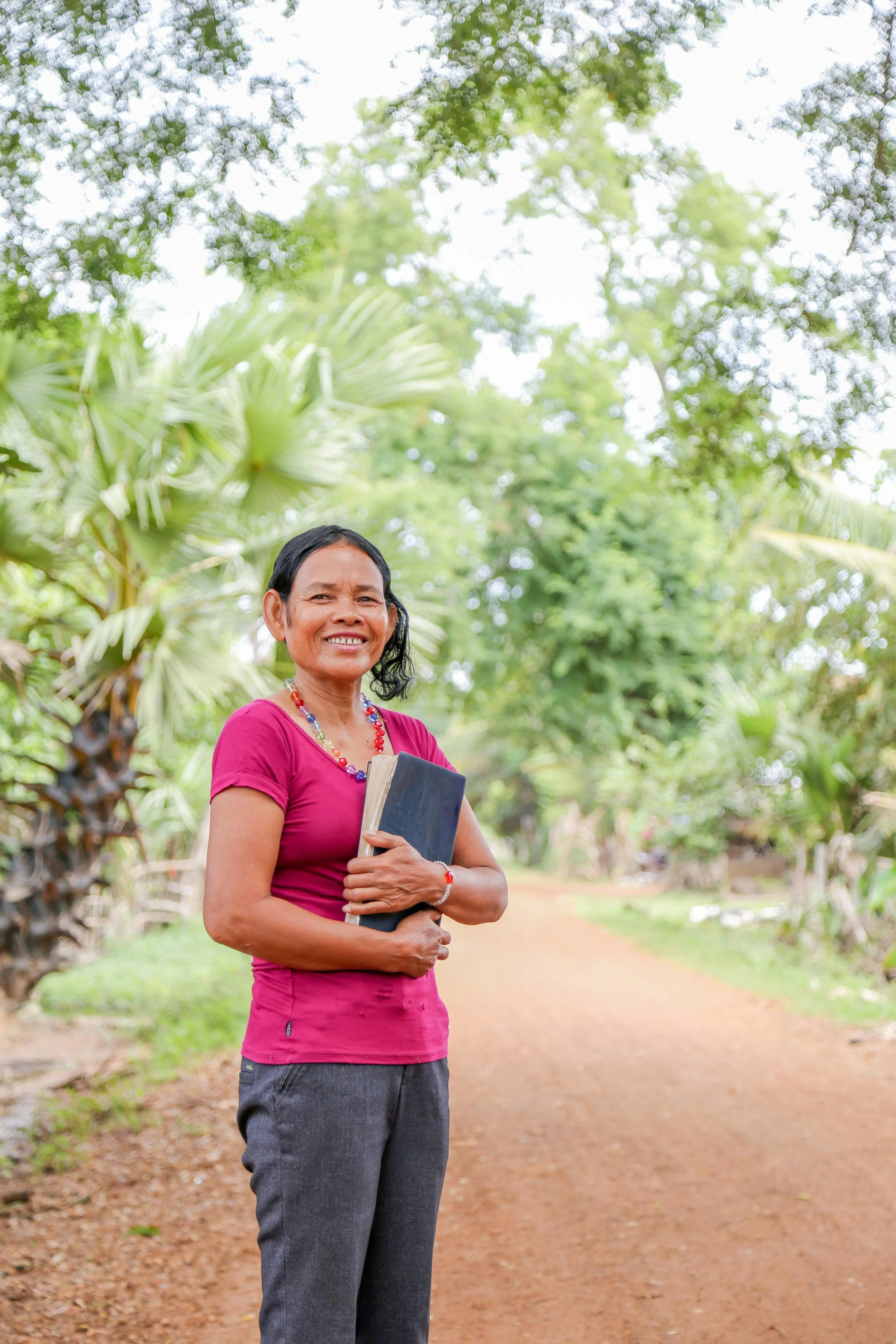 A smiling woman in a pink shirt and gray pants holding a book stands on a dirt path in a lush green outdoor setting.