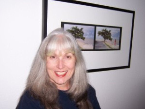 A smiling woman with long gray hair standing in front of two framed landscape photos on a white wall.