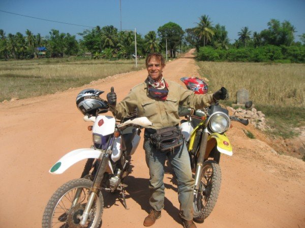 Man standing next to a dual-sport motorcycle on a dirt road in a rural area with trees and fields in the background, giving a thumbs-up gesture.
