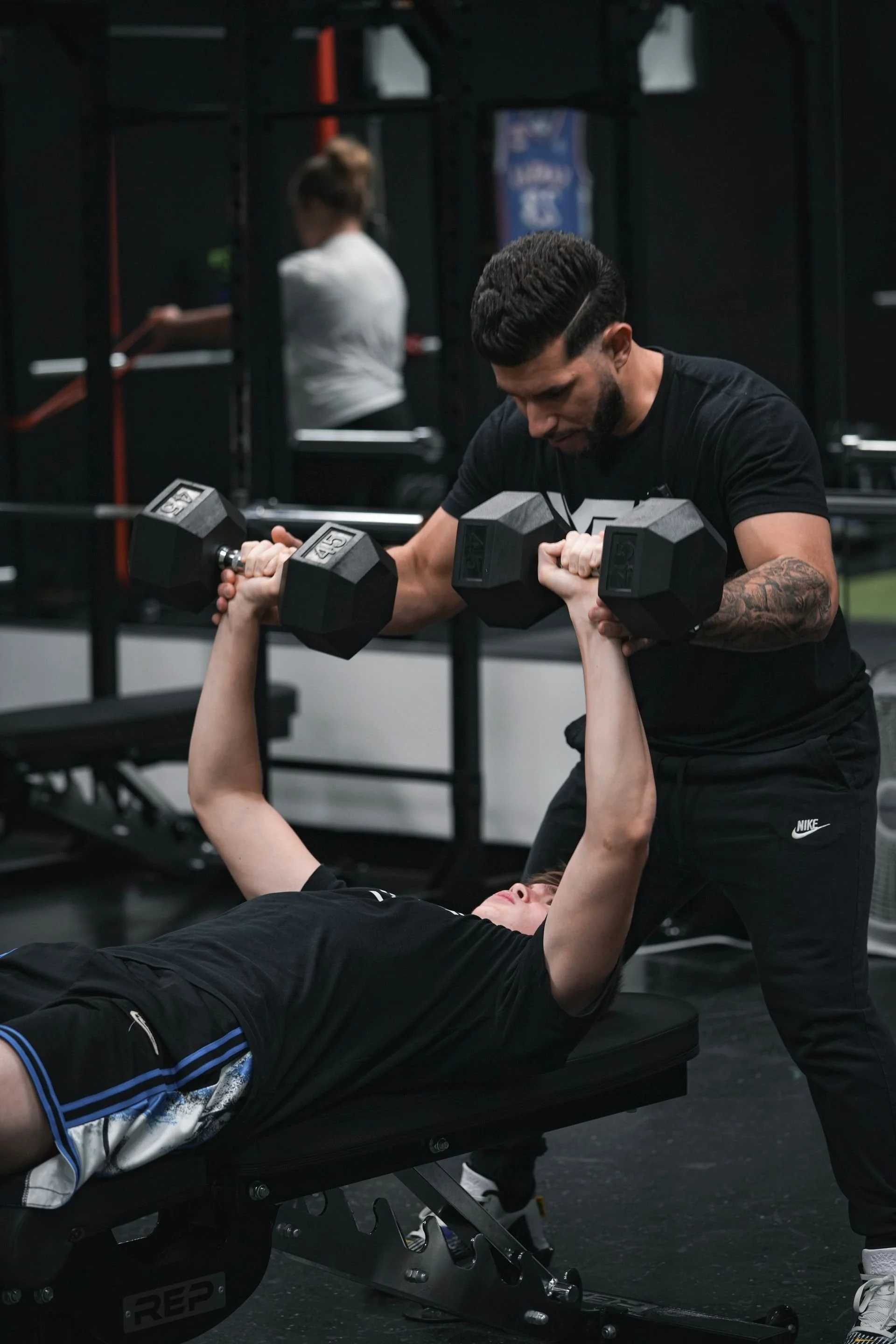 A fitness trainer assists a person lying on a bench press during workout in a gym, lifting dumbbells and focusing; gym equipment and a person in the background.