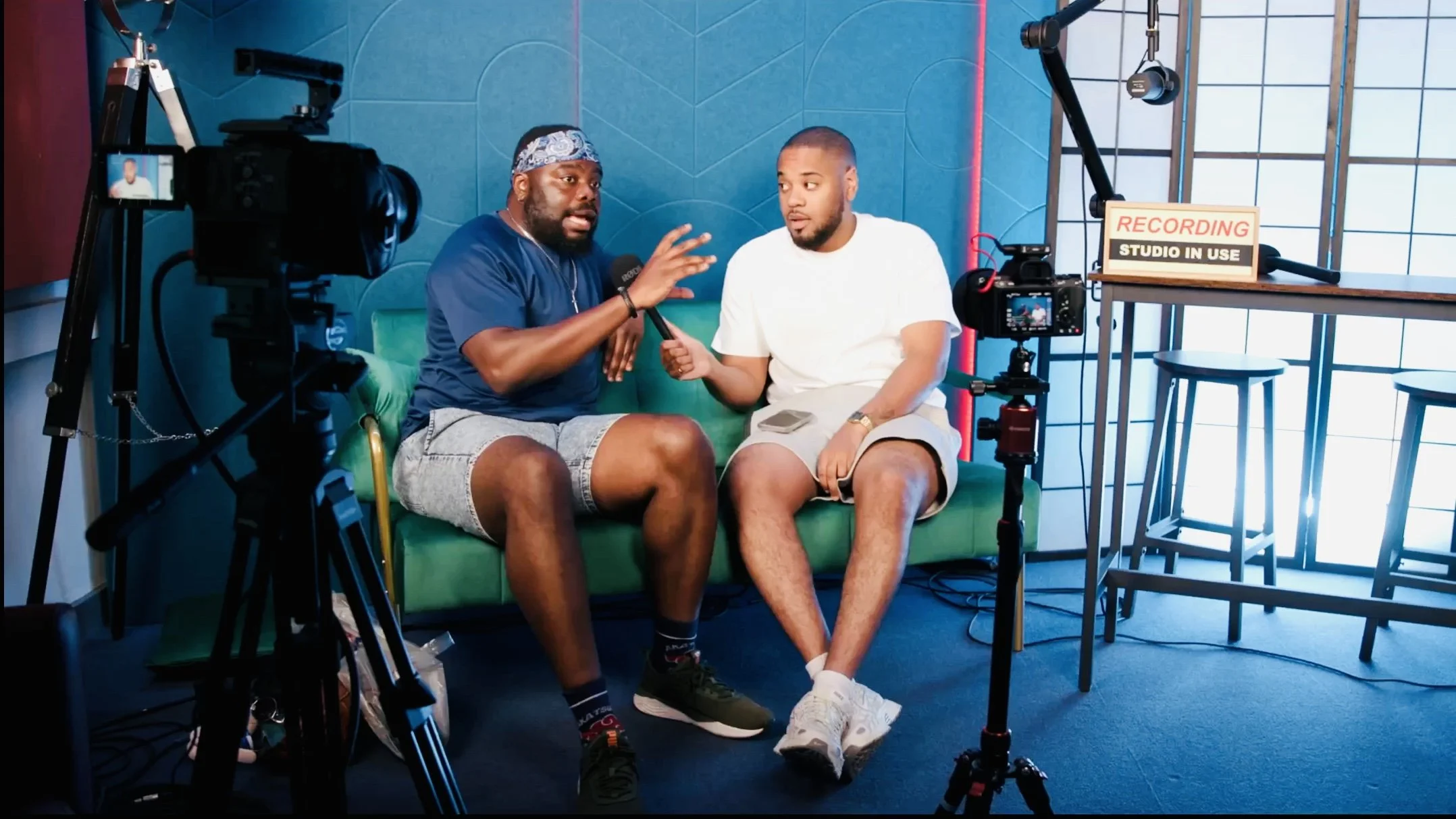 Two men seated on a green sofa in front of a blue wall during an interview or podcast recording. One man is speaking into a microphone, gesturing with his hand, while the other man listens. There is professional camera equipment set up around them, i