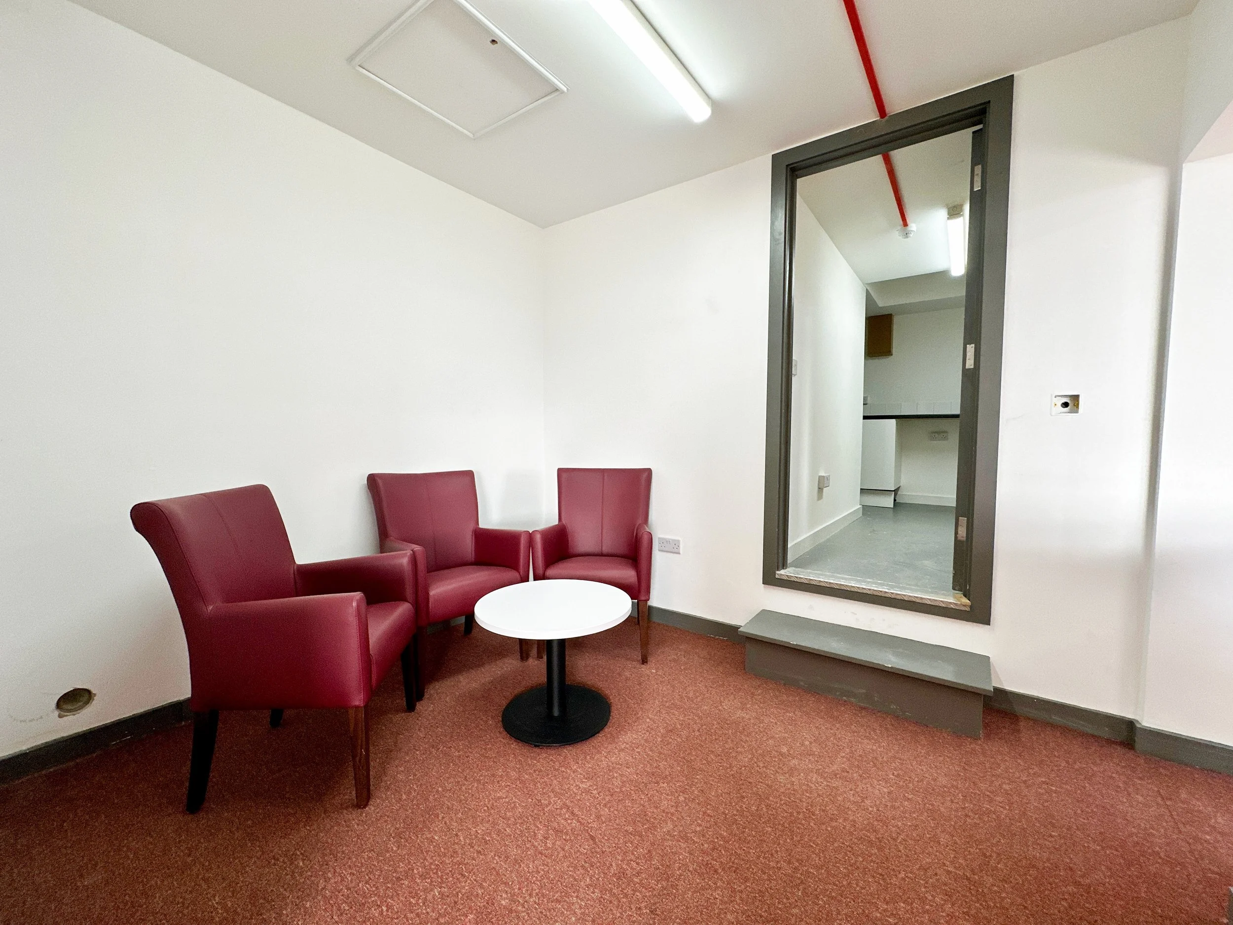 A small, furnished room with three red armchairs and a round white table, situated on a red carpet. There's a rectangular wall opening with a step leading to an adjacent room.