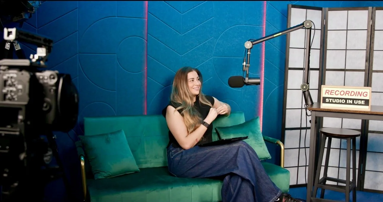 A woman in a recording studio sitting on a green velvet couch with pillows, smiling, with a microphone overhead, a camera on the left, and a sign that says 'Recording Studio in Use' on a table to the right.