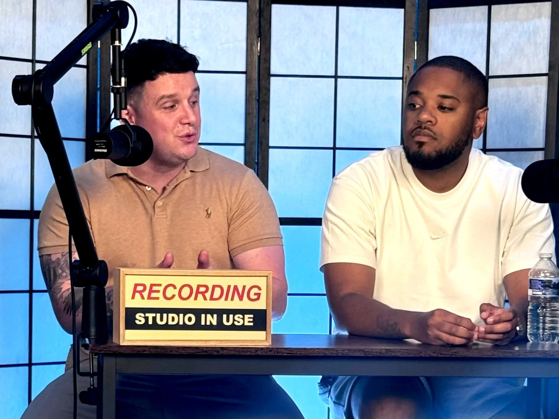 Two men seated at a table in a recording studio, with a sign that reads 'RECORDING STUDIO IN USE' in front of them, and microphones in front of each person.