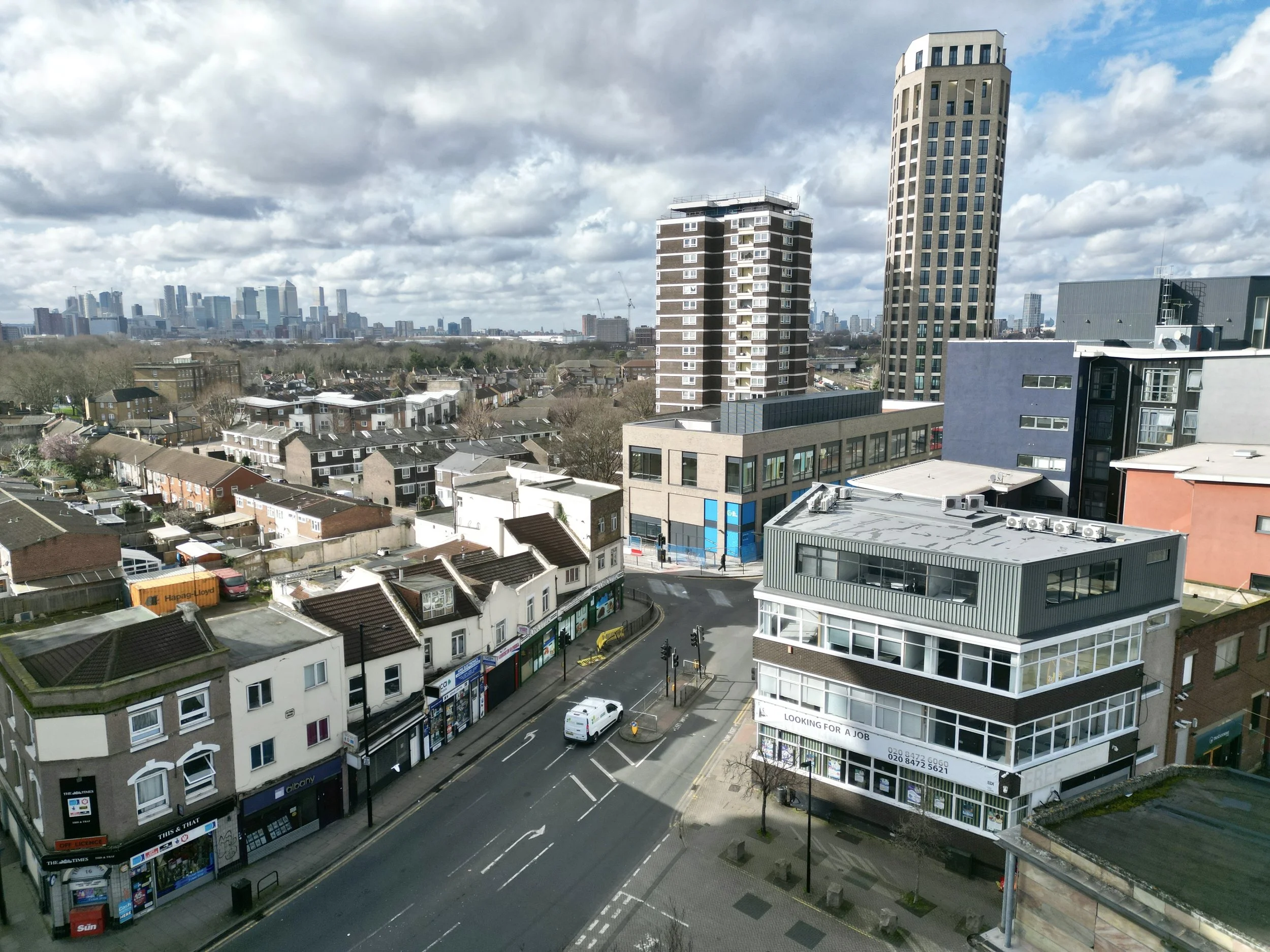 Aerial view of an urban area with a mix of residential and commercial buildings, including high-rises and smaller structures. A street with shops and a roundabout is visible, and a city skyline is in the distance under cloudy skies.