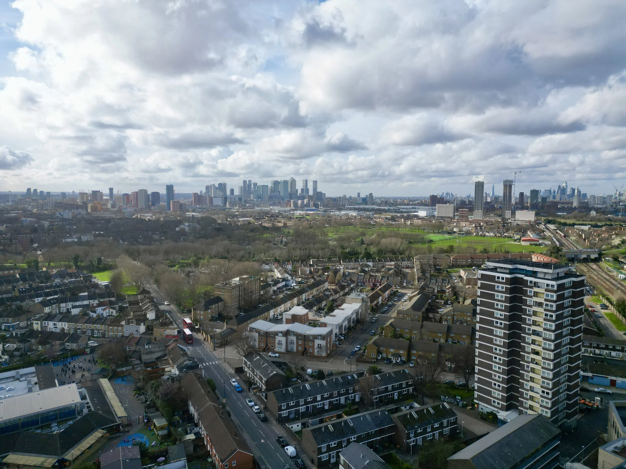 Aerial view of a cityscape with residential buildings, parks, and a skyline with skyscrapers in the background under a cloudy sky.