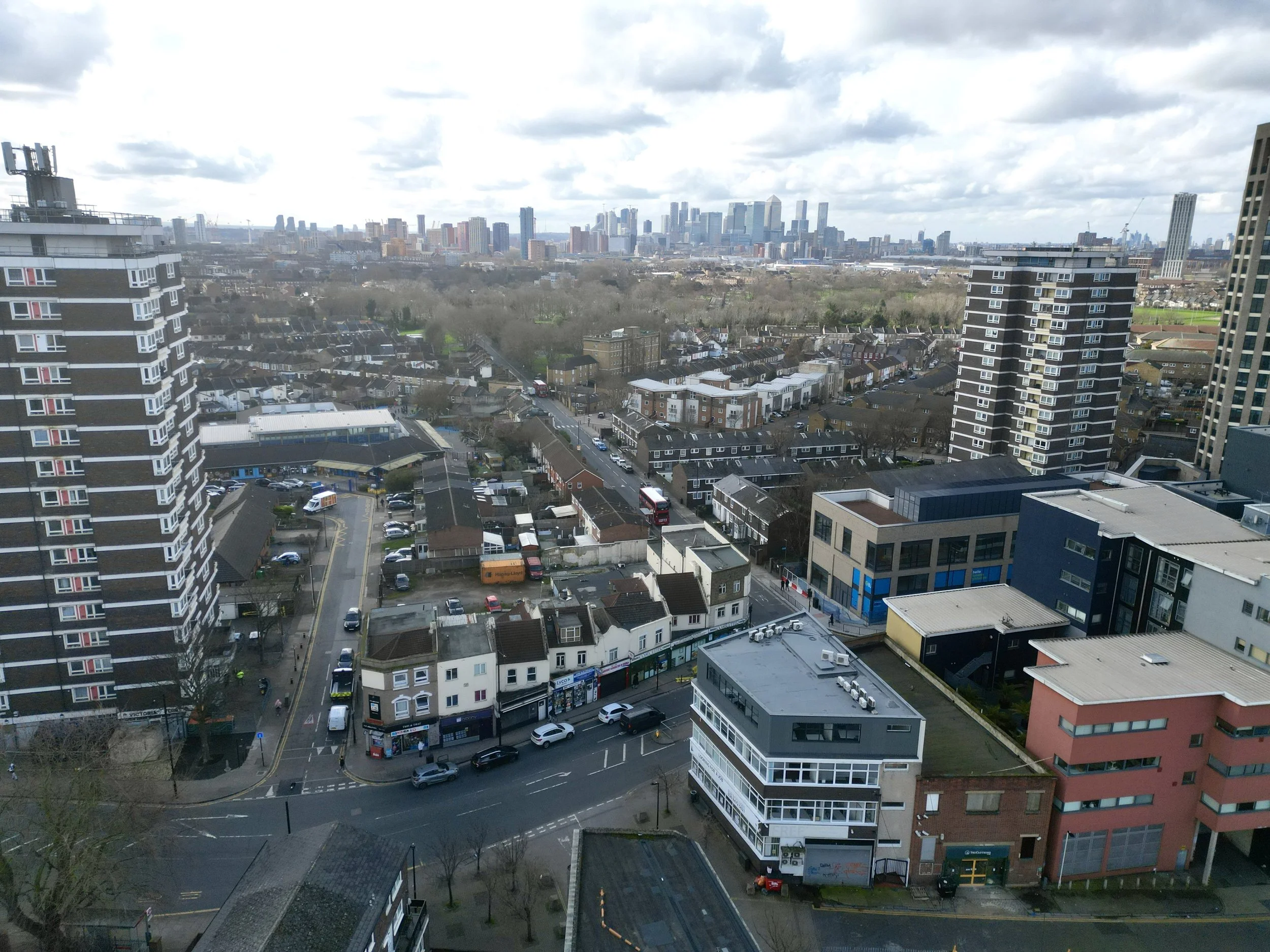 Aerial view of an urban neighborhood with high-rise buildings, streets, and parked cars, with a city skyline visible in the background under cloudy skies.