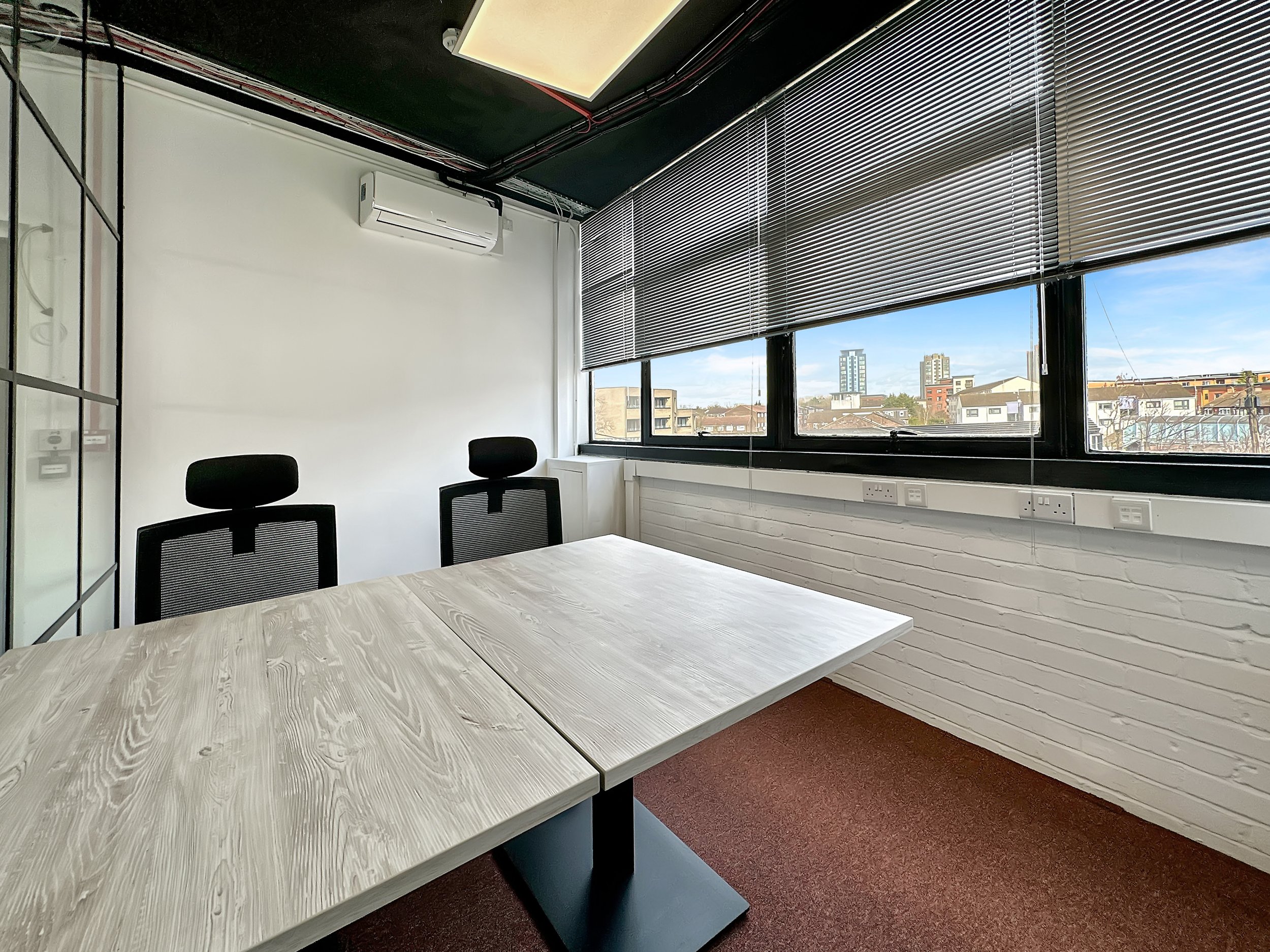 Minimalist office space with a wooden table, two black chairs, and large windows with blinds. City view and air conditioning unit visible.