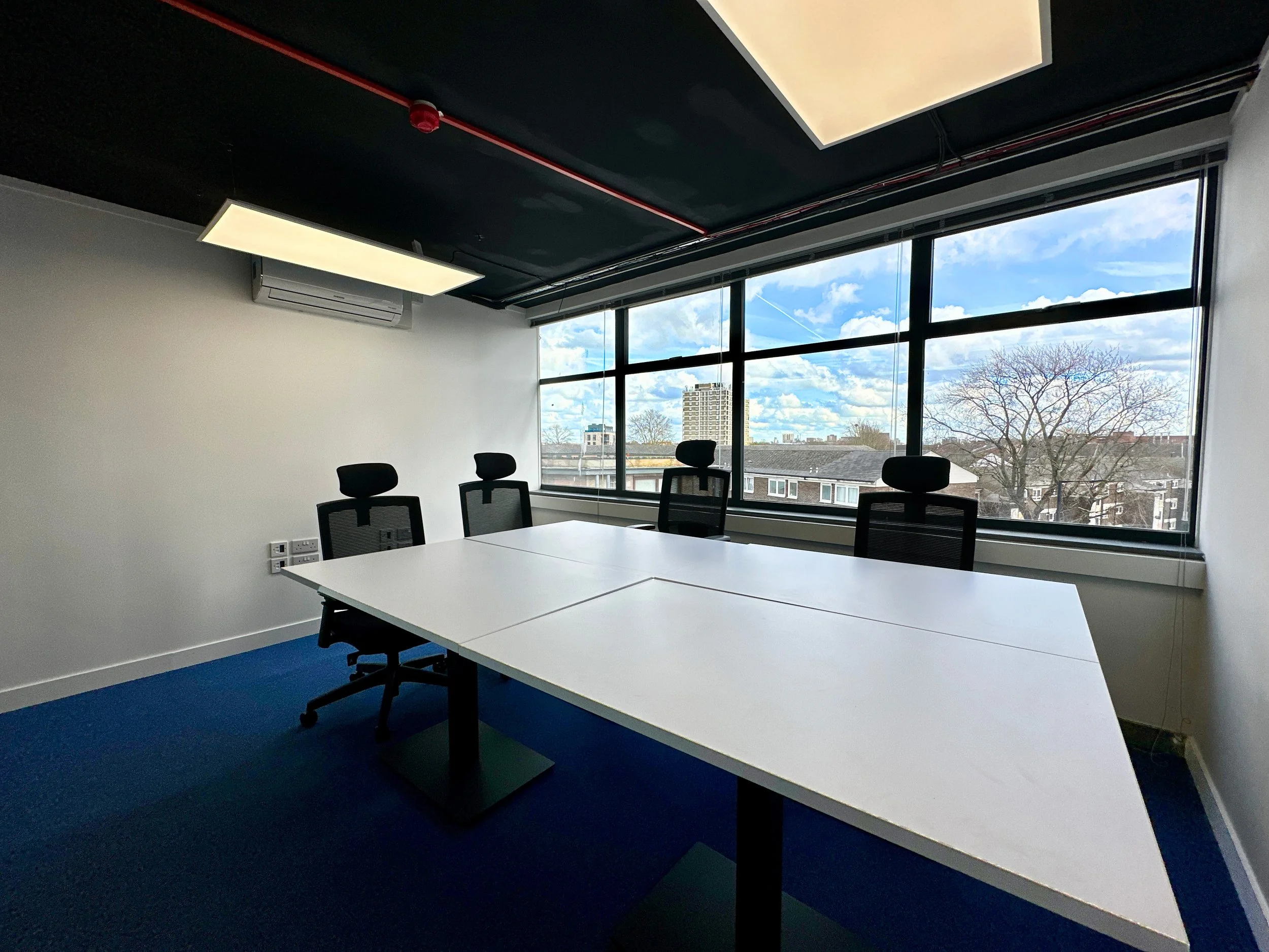 Modern office conference room with a large white table, four black office chairs, and a large window offering a city view. Overhead lighting and an air conditioning unit are present. Room has blue carpet and white walls.