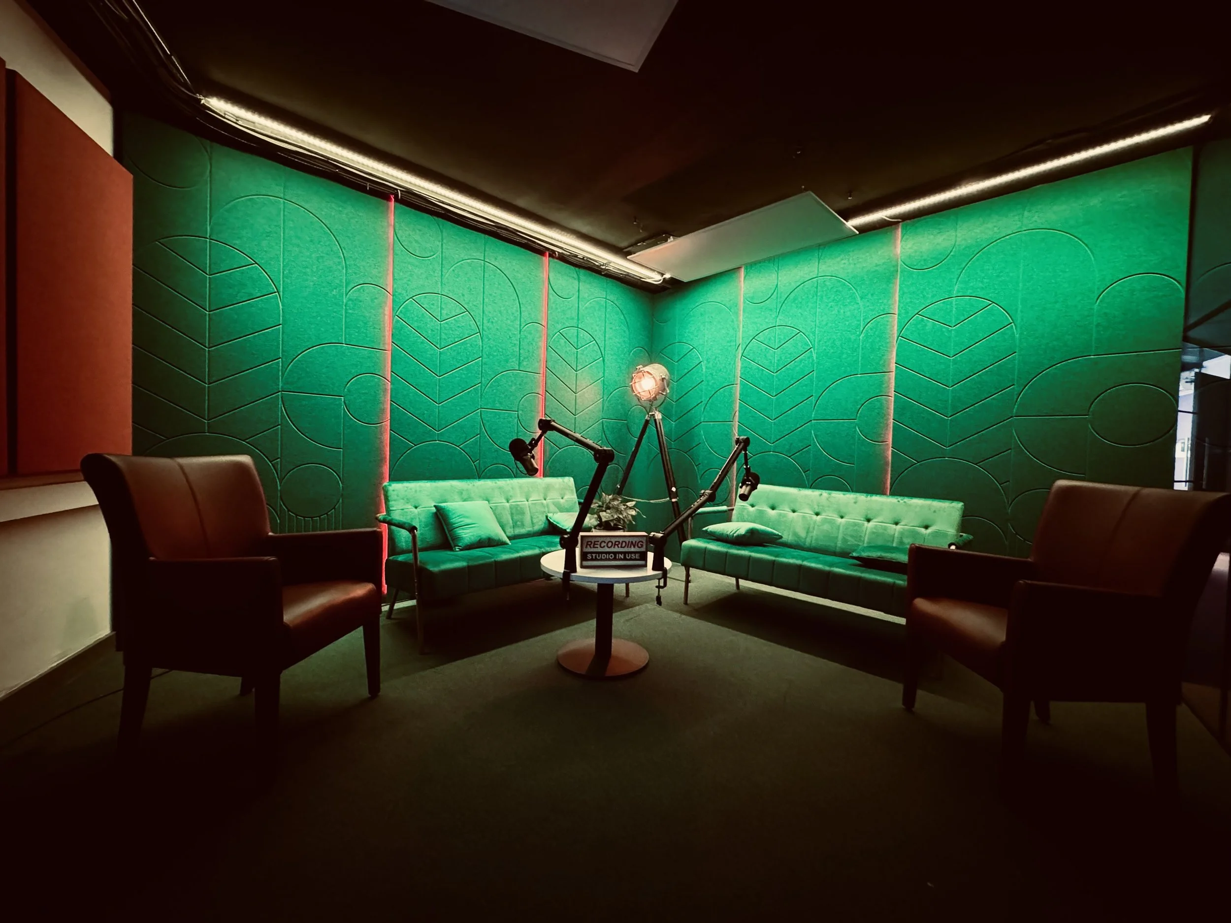 Recording studio with two green sofas and two brown armchairs arranged in a semi-circle. A small round table in the middle holds a microphone and a sign that says 'RECORDING STUDIO IN USE'. The background features a green wall with geometric patterns
