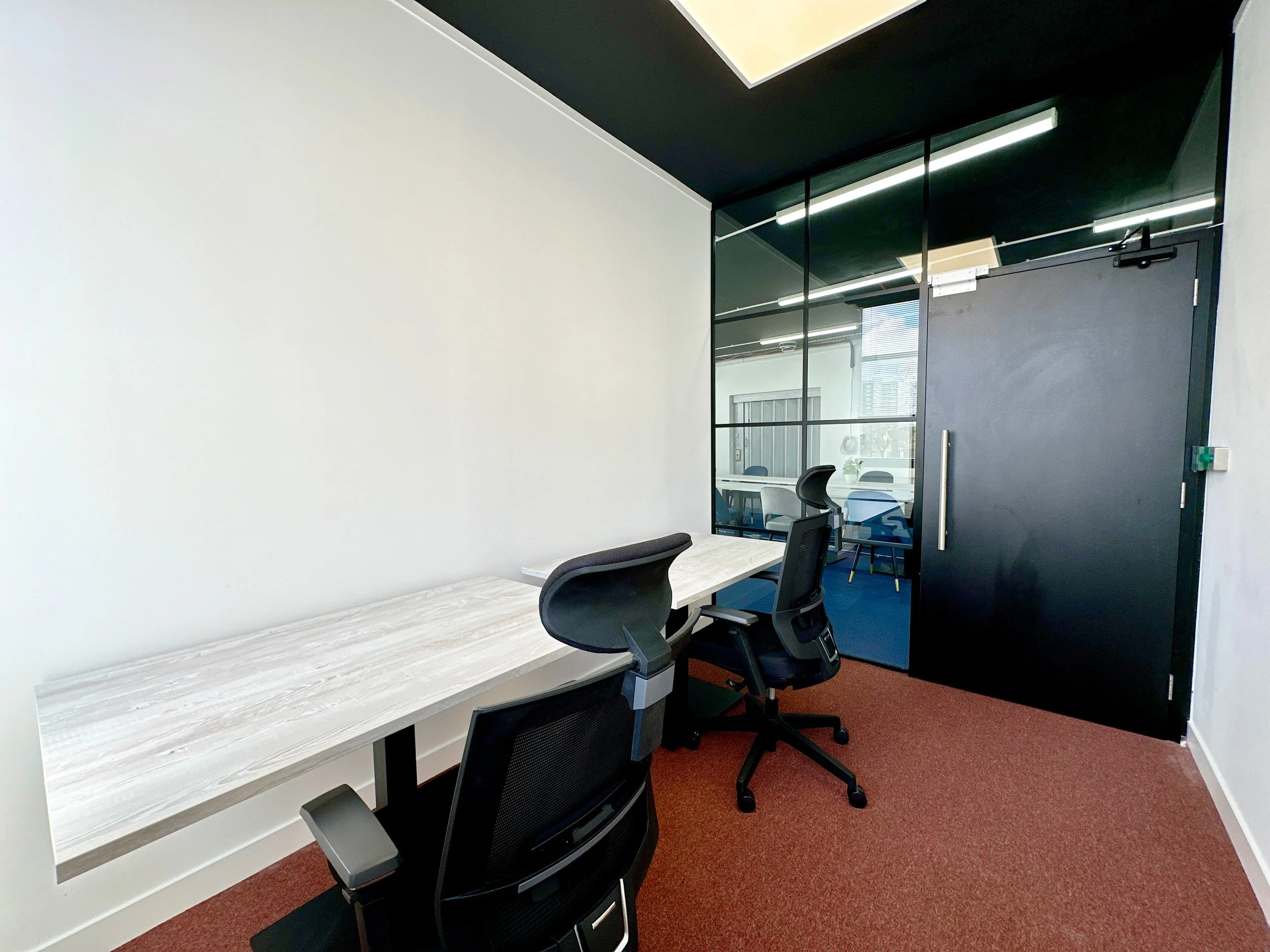 Small modern office space with two ergonomic chairs, a wooden desk, glass wall, and dark door. Carpeted floor, white walls, and ceiling lights.