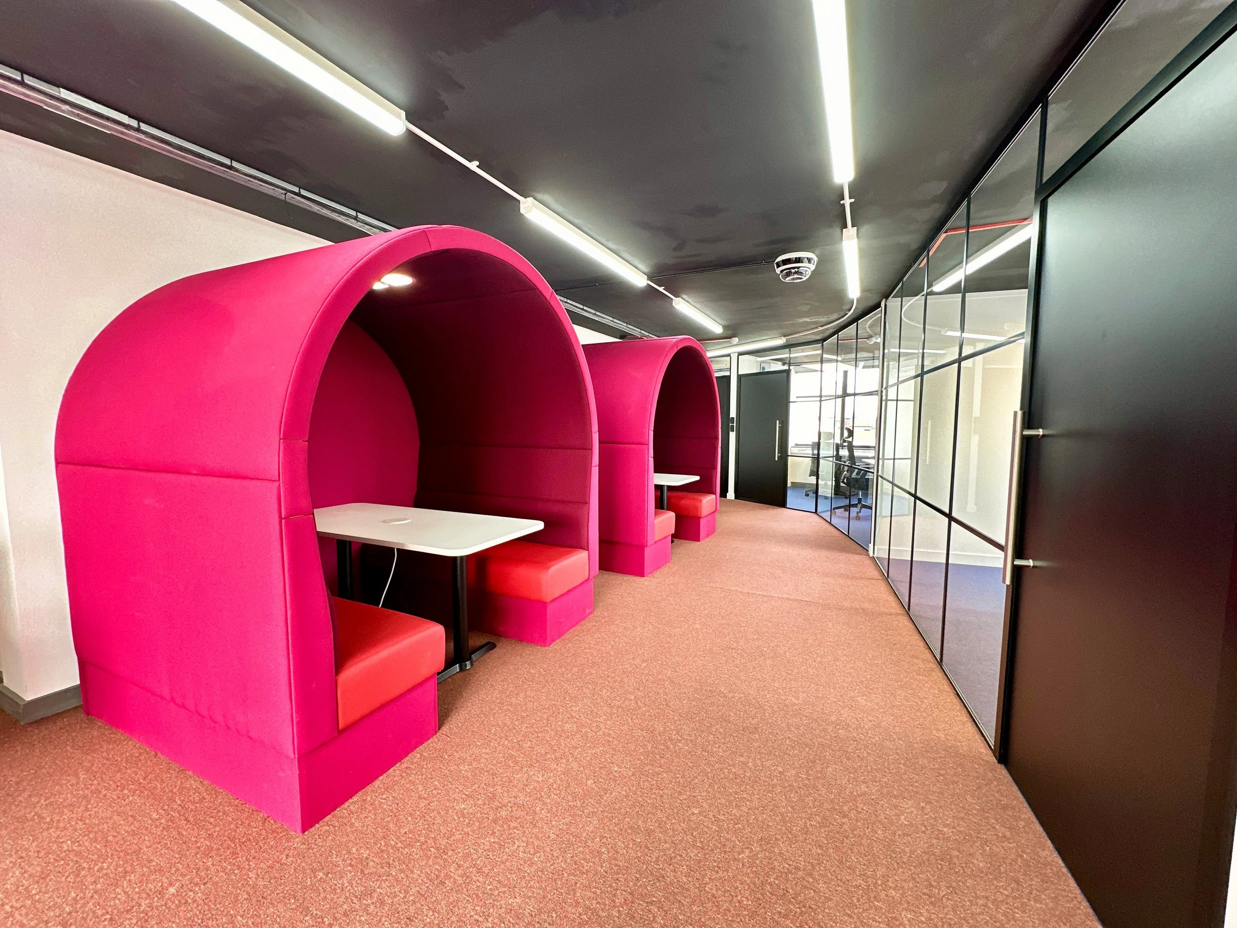 Modern office space with pink pod-like seating, white tables, and a glass-walled meeting room.